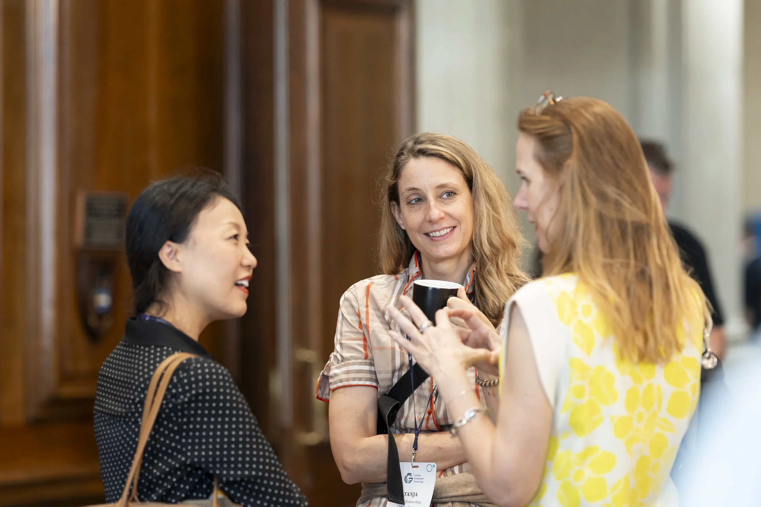 Three women engaged in conversation at an indoor event. The woman in the middle is holding a black cup and smiling, wearing a checked shirt and a conference badge. The woman on the right, with long red hair and glasses on her head, is wearing a yellow patterned dress. The woman on the left, with dark hair, is wearing a black and white polka dot top, carrying a tan purse.