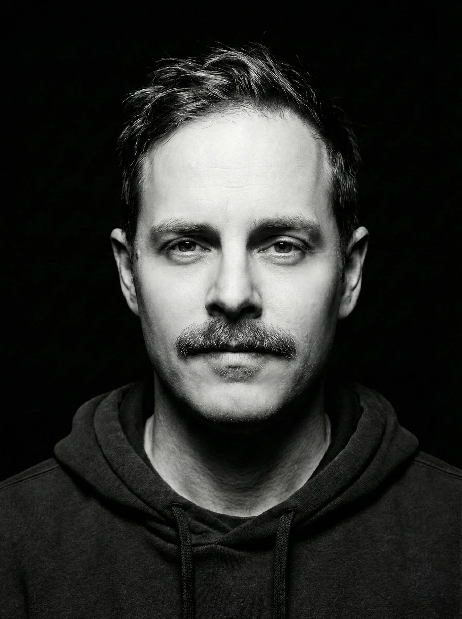 Black and white portrait of a man with short hair and a mustache, wearing a hoodie, looking directly at the camera.