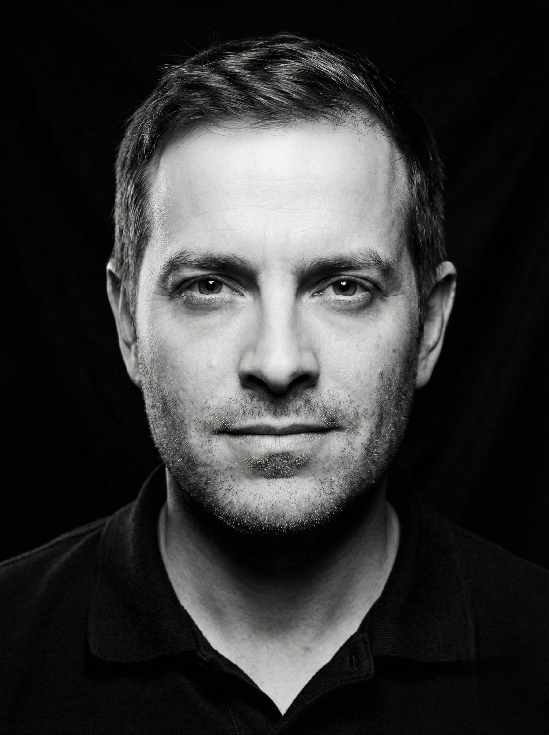 Black and white portrait of a man with short hair and facial hair, wearing a dark shirt, against a dark background.
