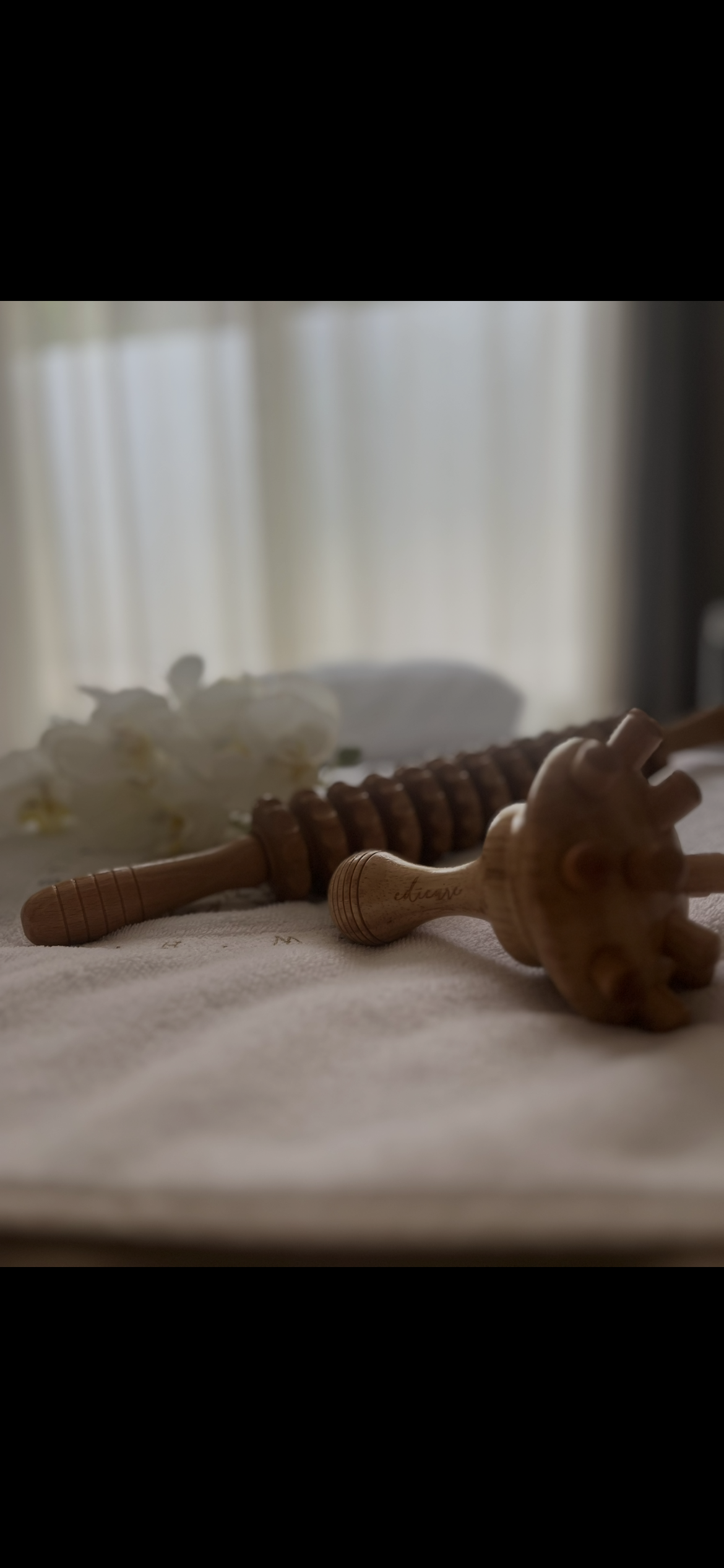 Deux masseurs en bois posés sur une table, avec des fleurs blanches floues en arrière-plan et une lumière naturelle provenant d'une fenêtre.