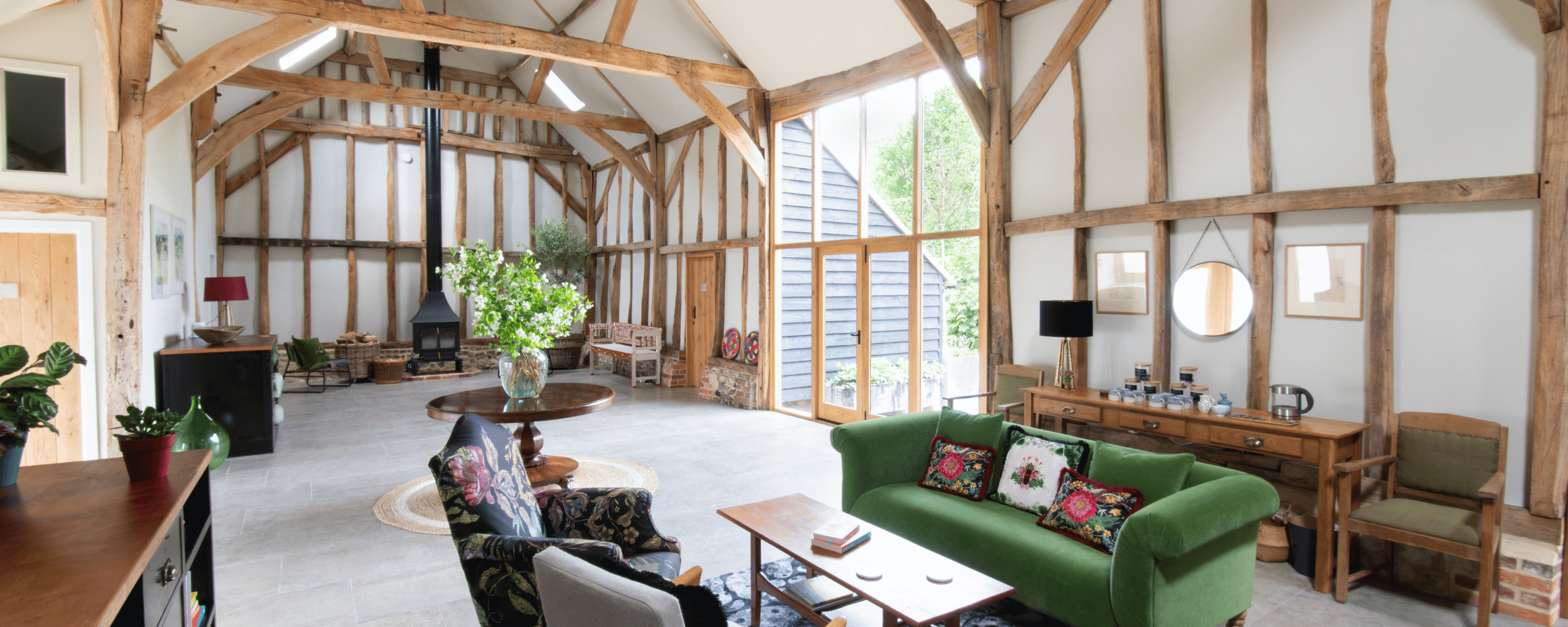 Interior of a rustic living room with exposed wooden beams, large windows, green sofa, floral armchair, and a fireplace with a black chimney.