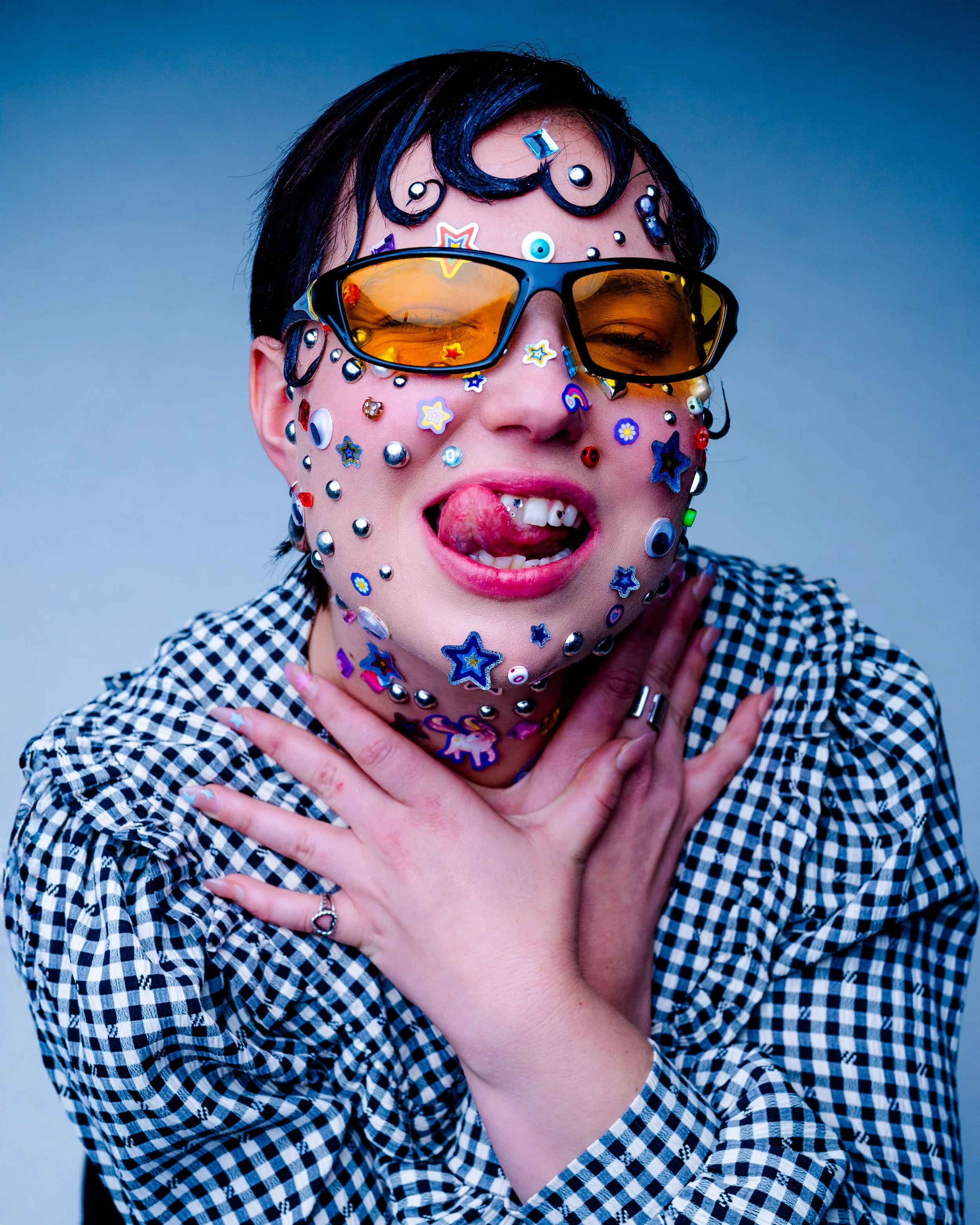 Person covered in colorful stickers, wearing sunglasses, sticking out tongue, and holding hands near neck against a gray background.