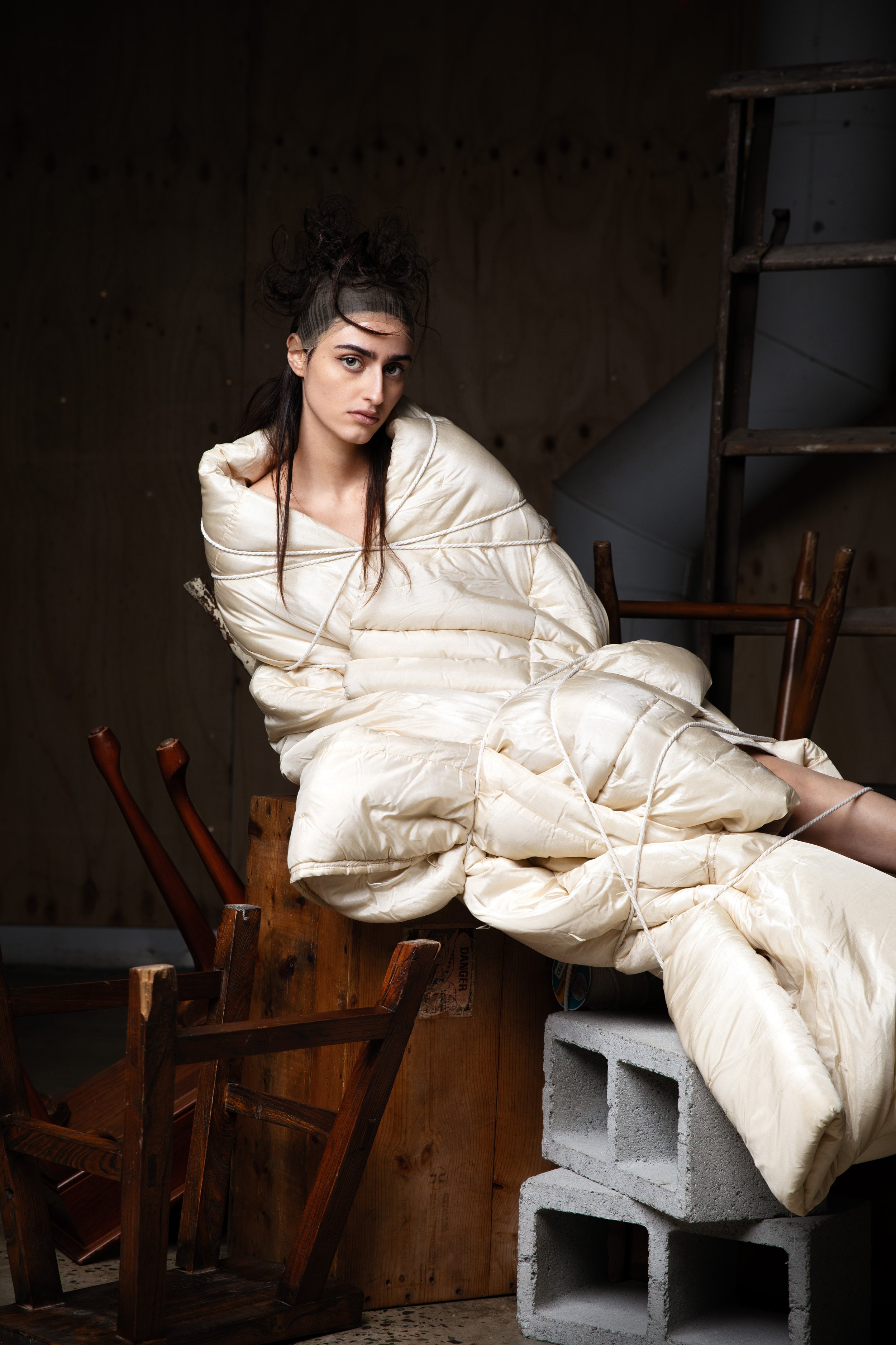 A woman wearing a puffy, off-white insulated suit, sitting on stacked cinder blocks and surrounded by overturned wooden chairs in a dimly lit, unfinished space.
