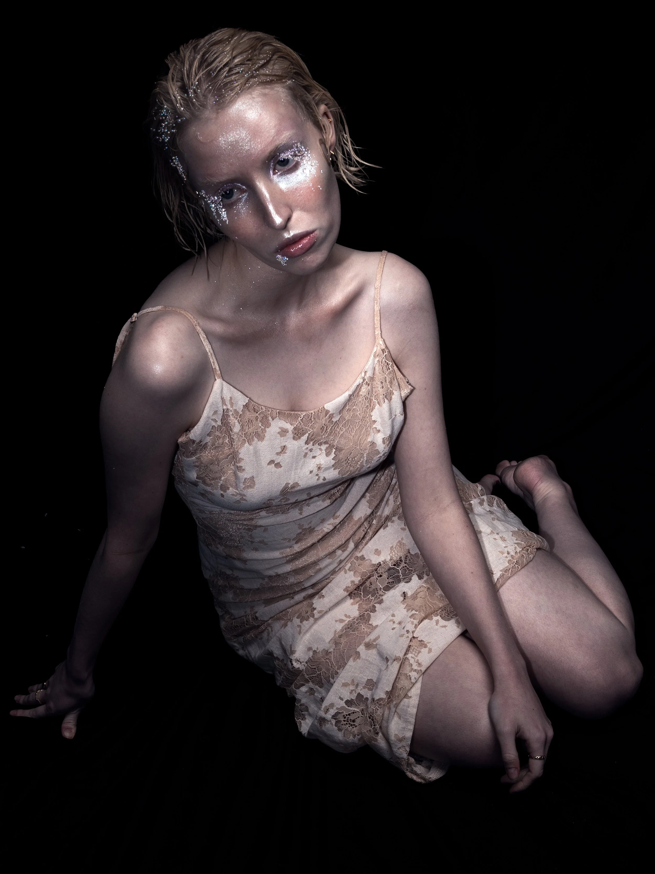 A woman with short, wet hair and glitter on her face, sitting on her heels against a black background, wearing a beige floral dress.