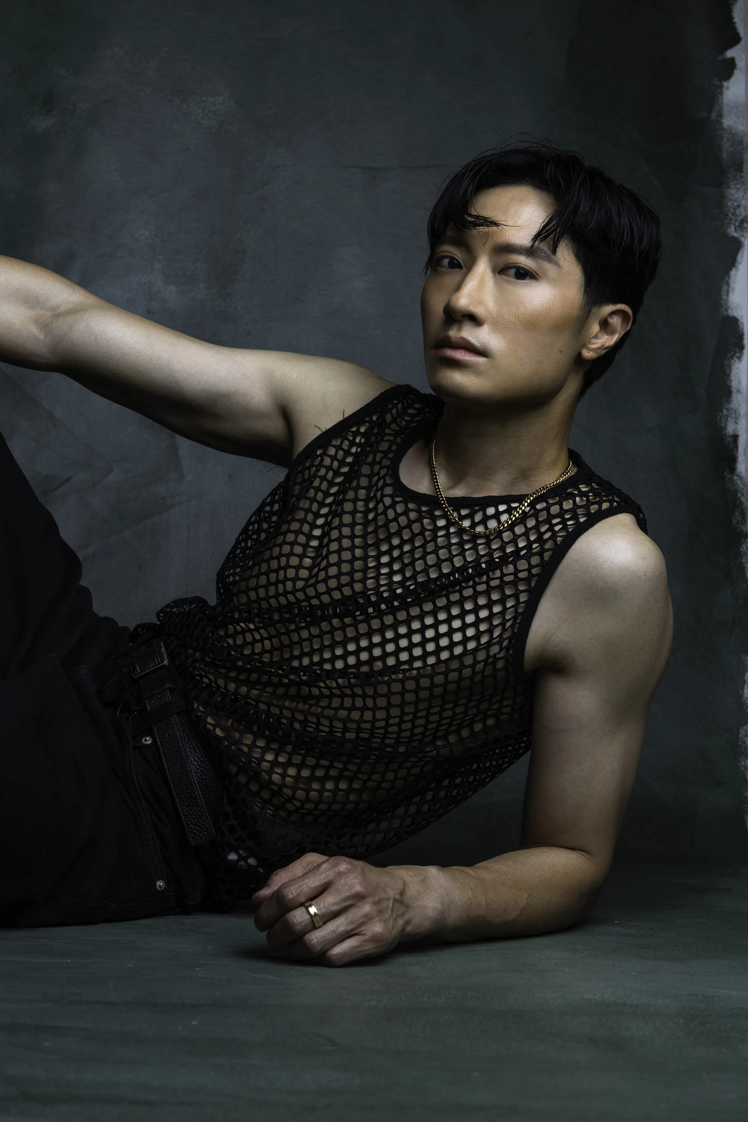 A young man with short black hair, wearing a black mesh sleeveless top and black pants, posed against a dark textured background.