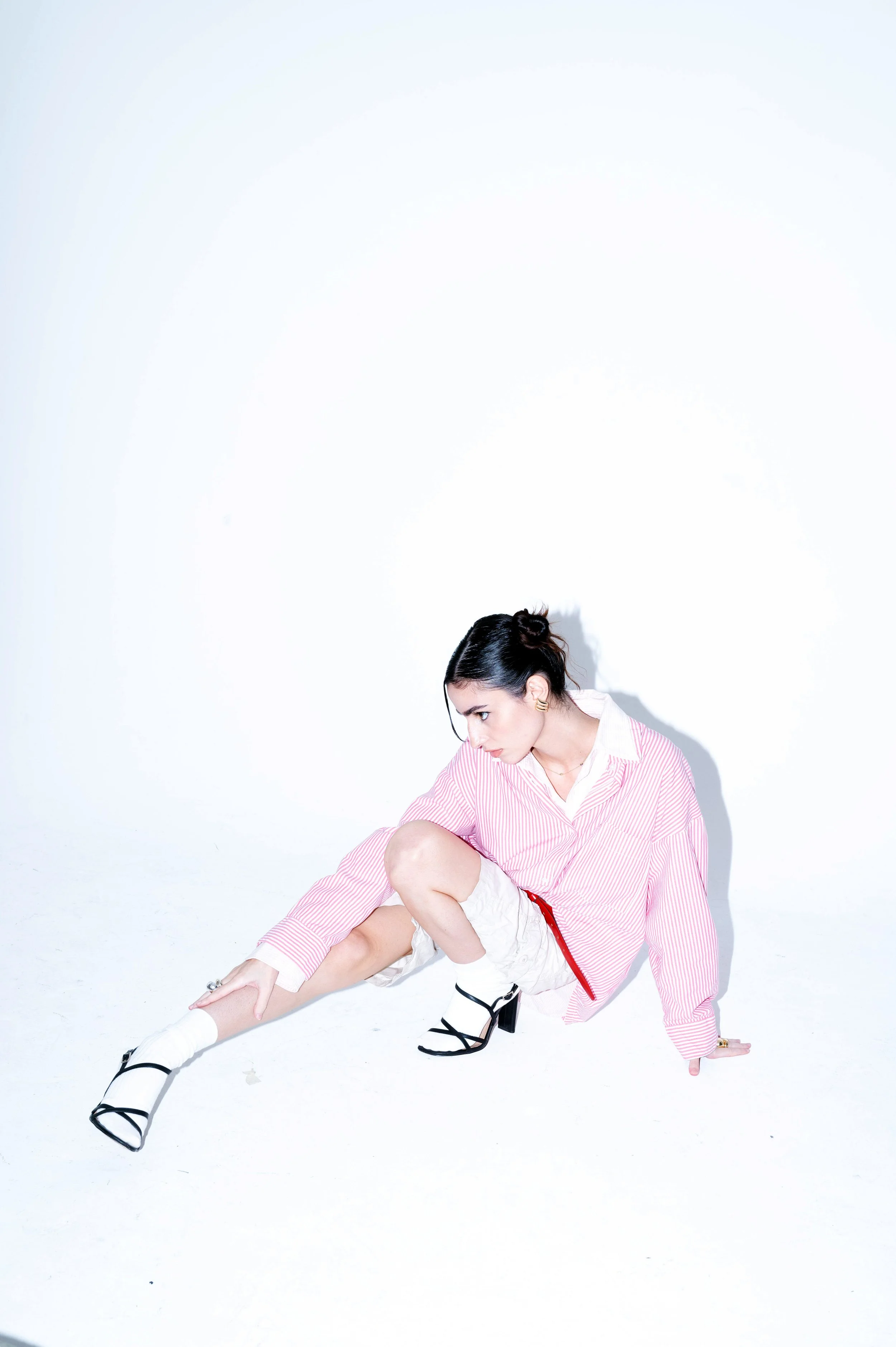 A woman crouching on the floor against a plain white background, wearing a pink and white striped shirt, beige shorts, and black high-heeled sandals.