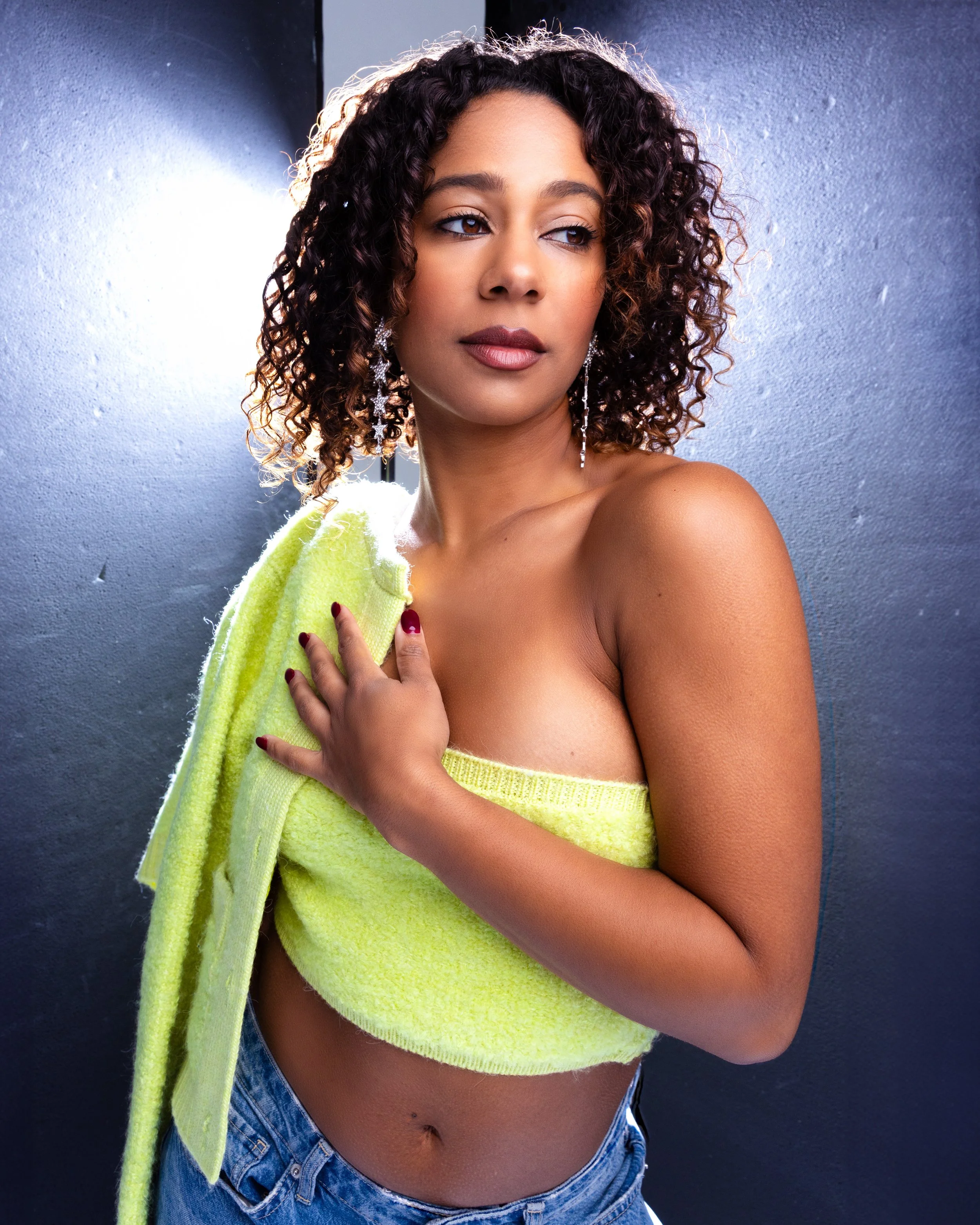 A woman with curly hair and earrings, wrapped in a yellow towel, stands against a dark gray background, looking to the side.