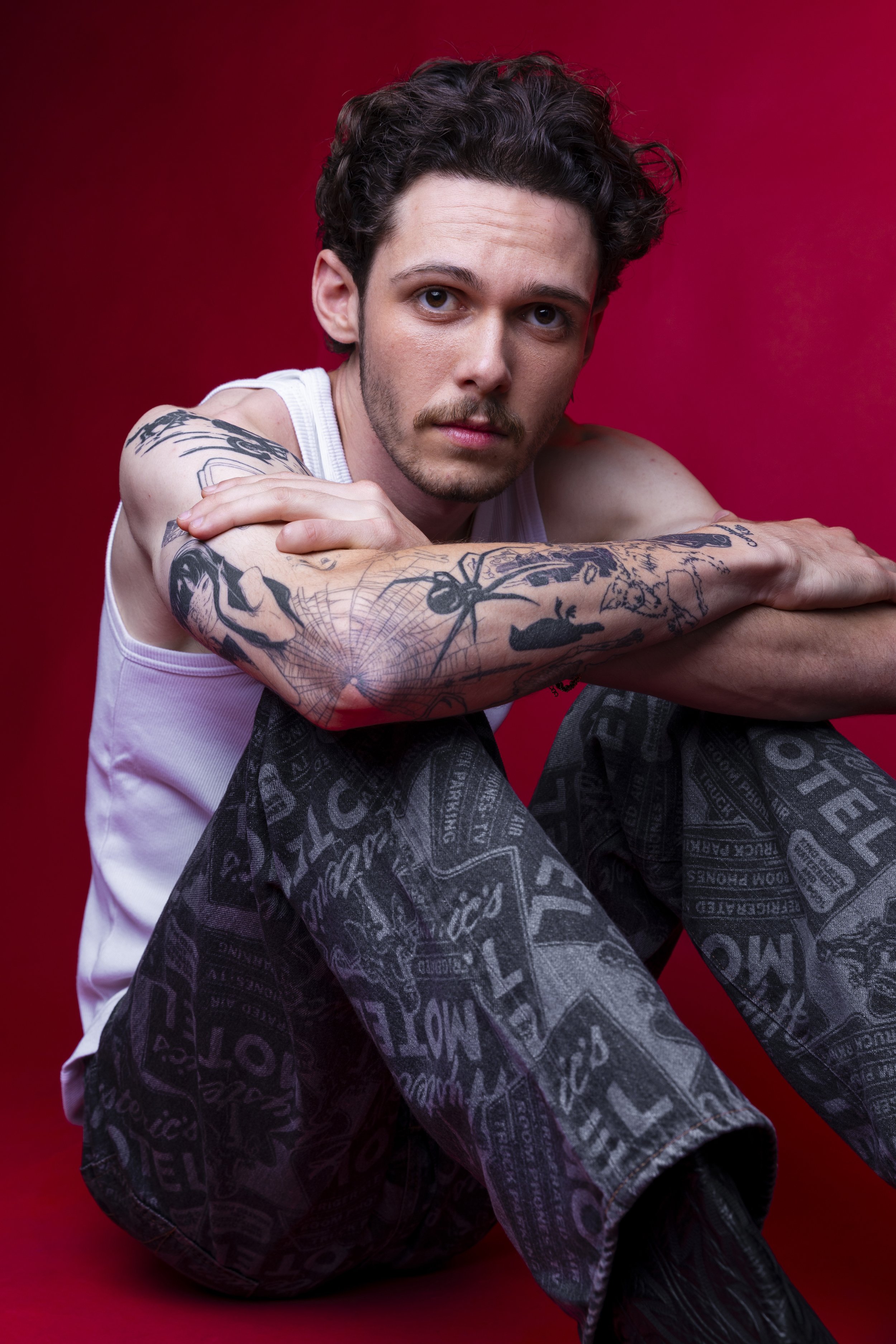 A young man with curly dark hair and tattoos, sitting against a red background, wearing a white tank top and black printed pants, looking at the camera.