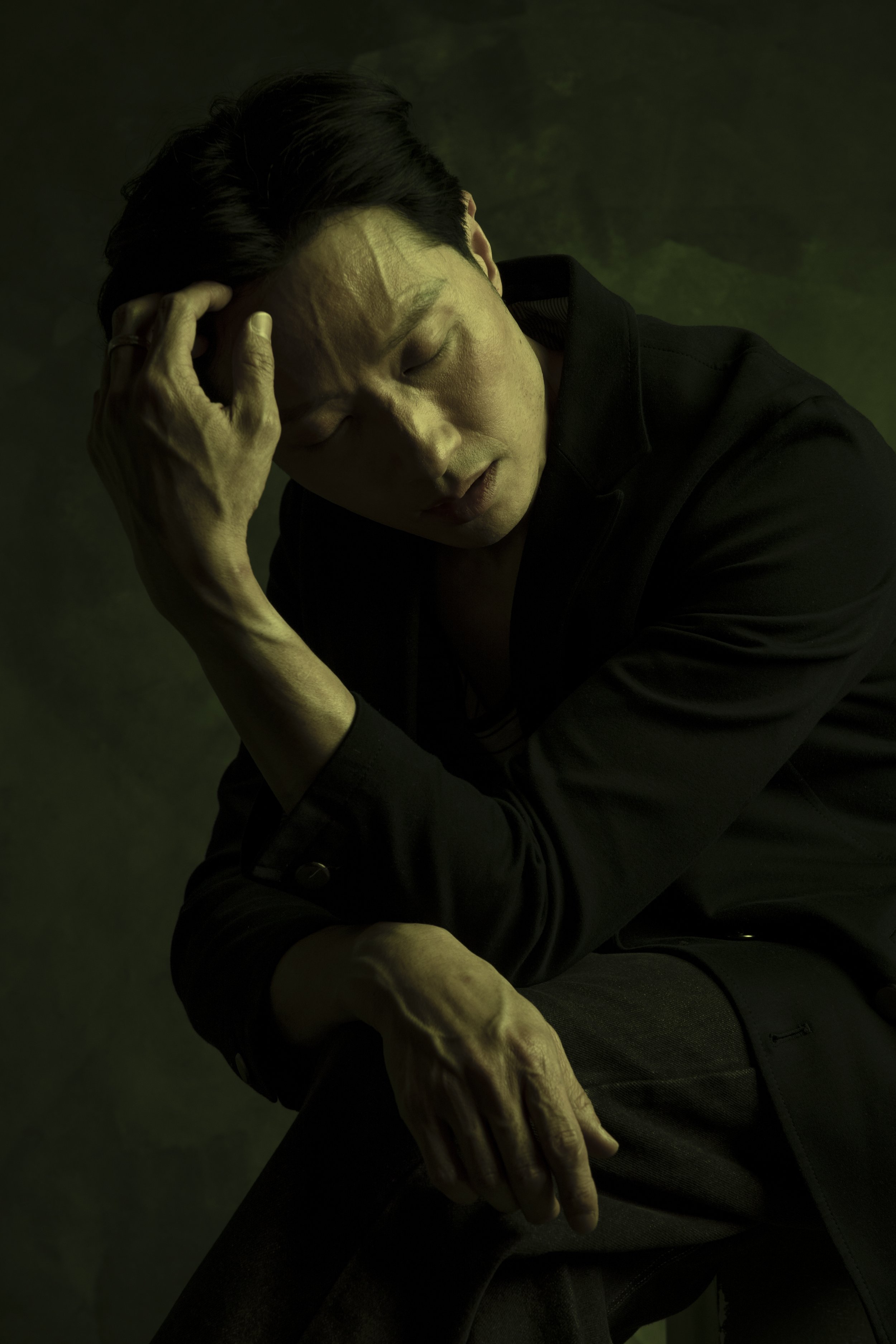 A man with dark hair in a black shirt, sitting with his eyes closed, holding his head with one hand, and tilting forward in a contemplative pose.