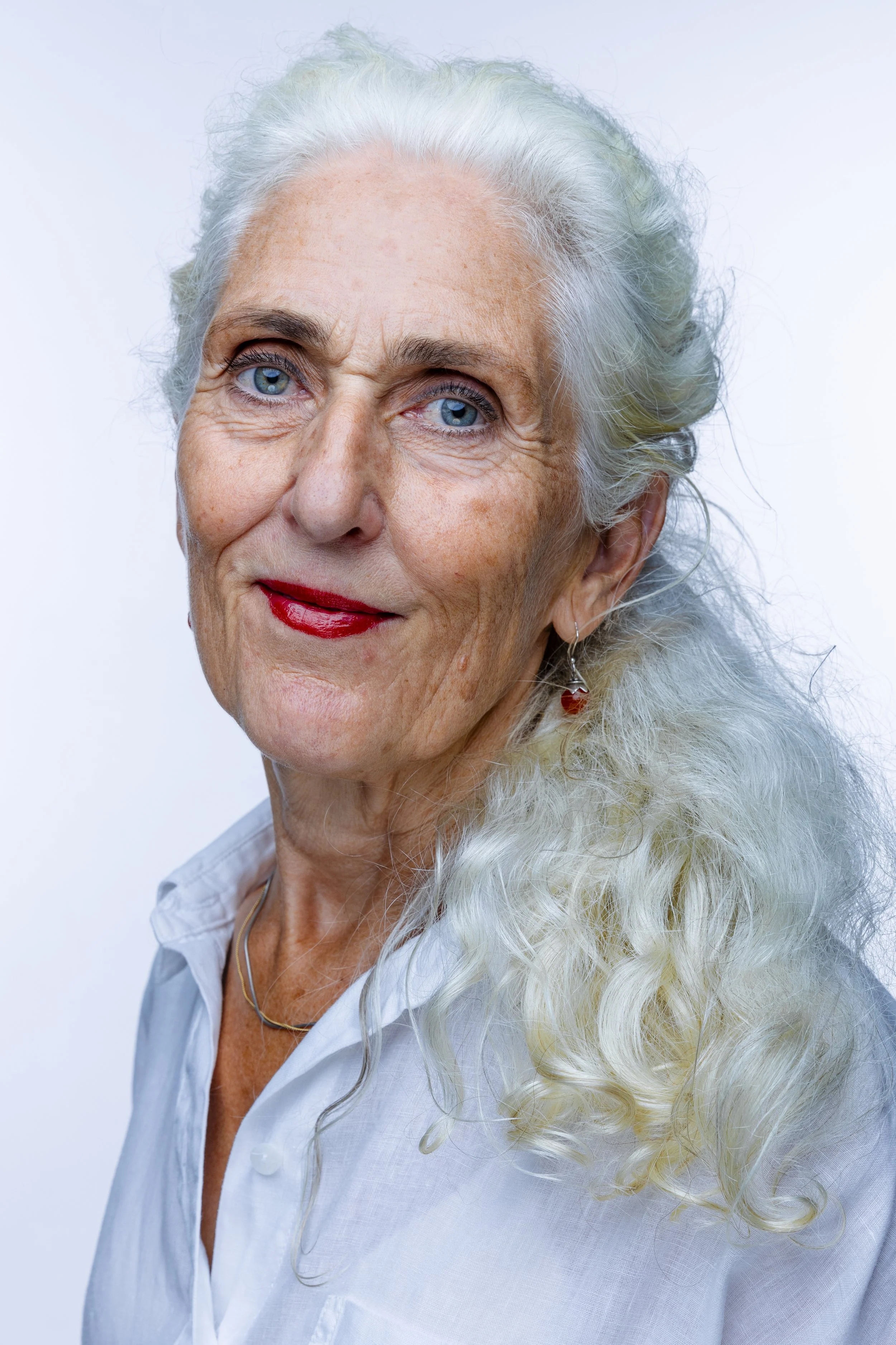 Close-up portrait of an elderly woman with long, curly white hair, blue eyes, and red lipstick, wearing a white shirt and earrings, against a plain light background.