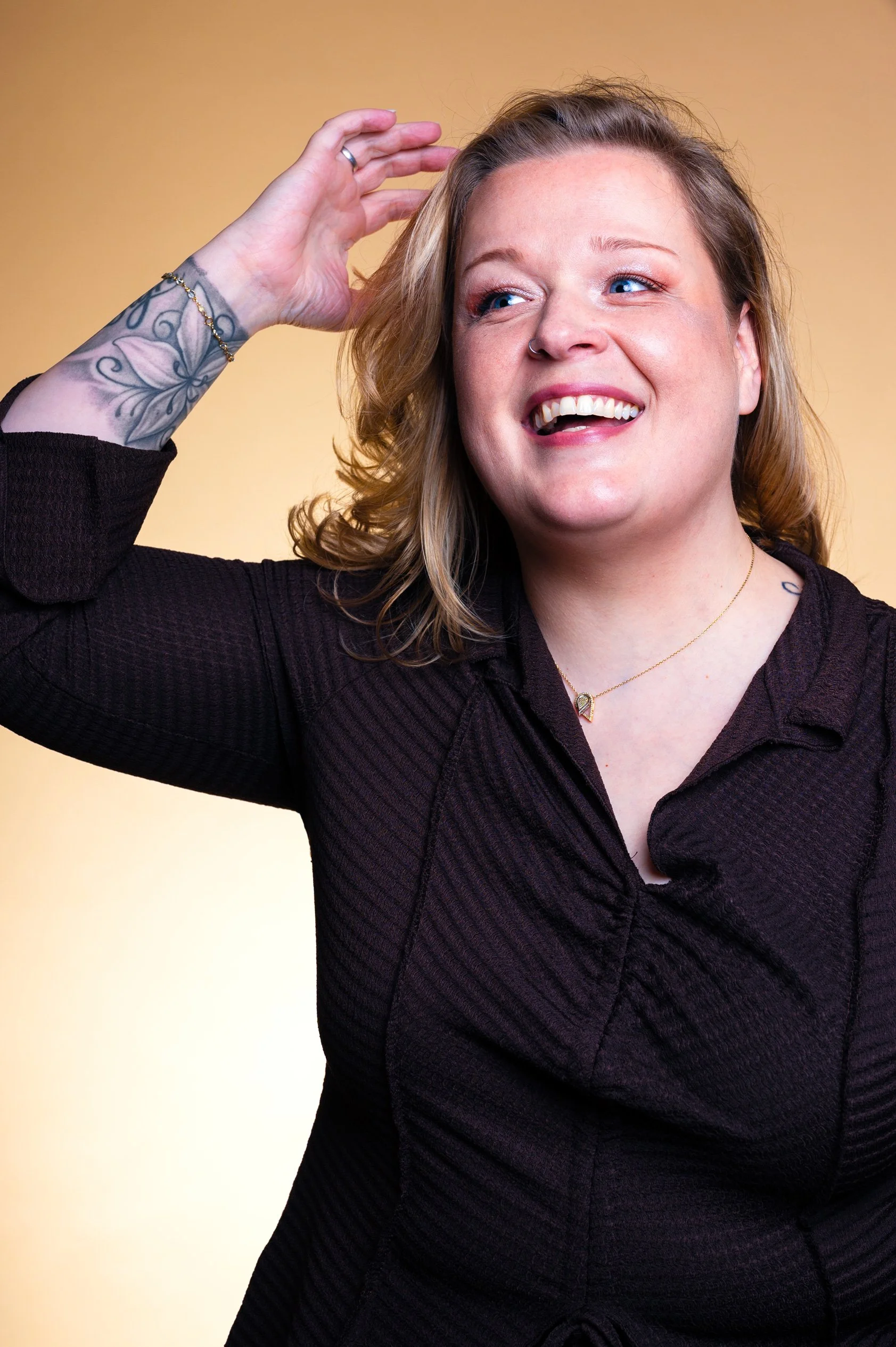 A smiling woman with blonde hair, a nose ring, and tattoos on her left arm poses against a yellow background.
