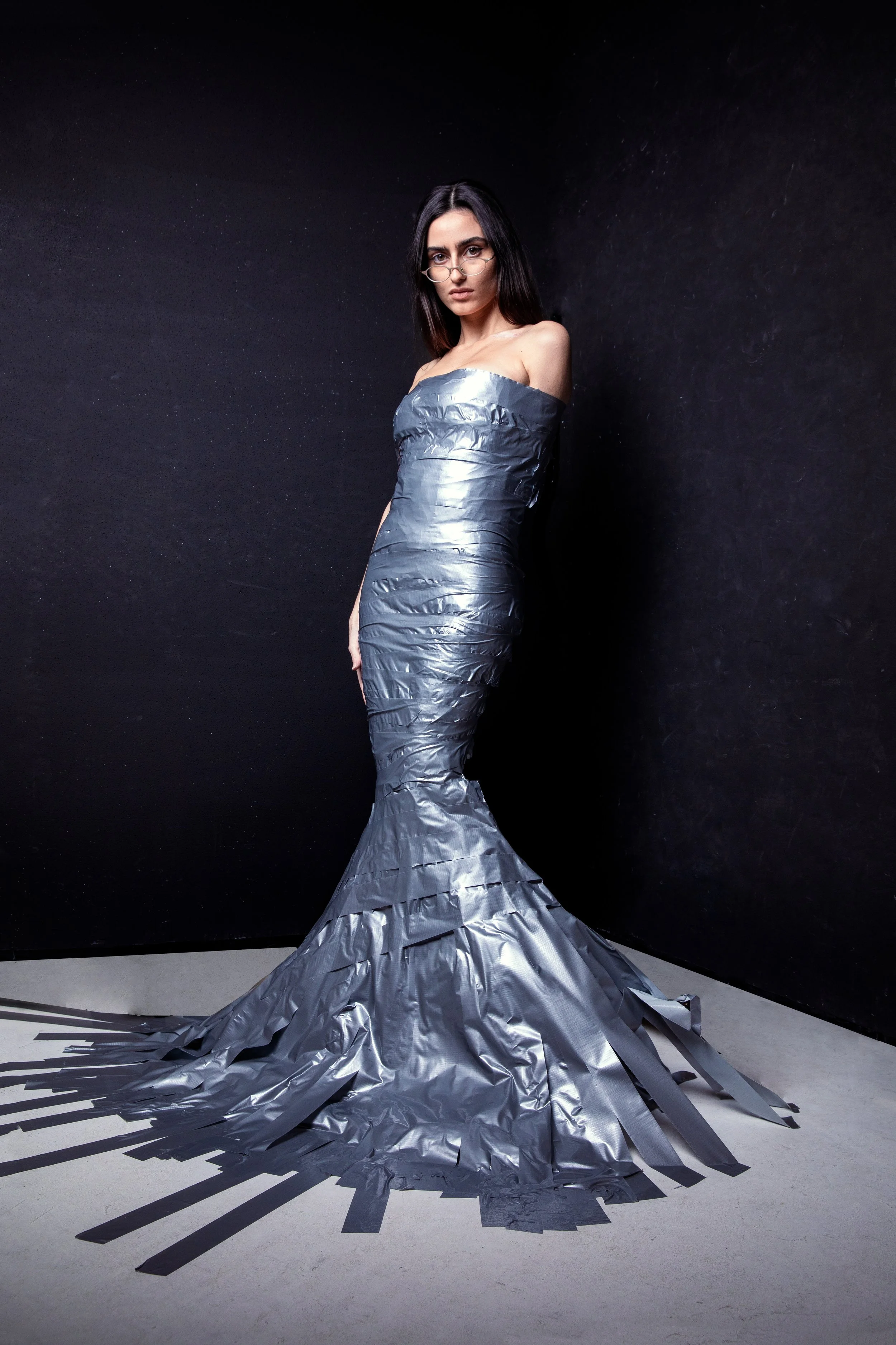 A woman with long dark hair and glasses wearing a silver metallic gown that drapes down to the floor, with a mermaid style and a train spreading out on the floor in a minimalist black and gray setting.