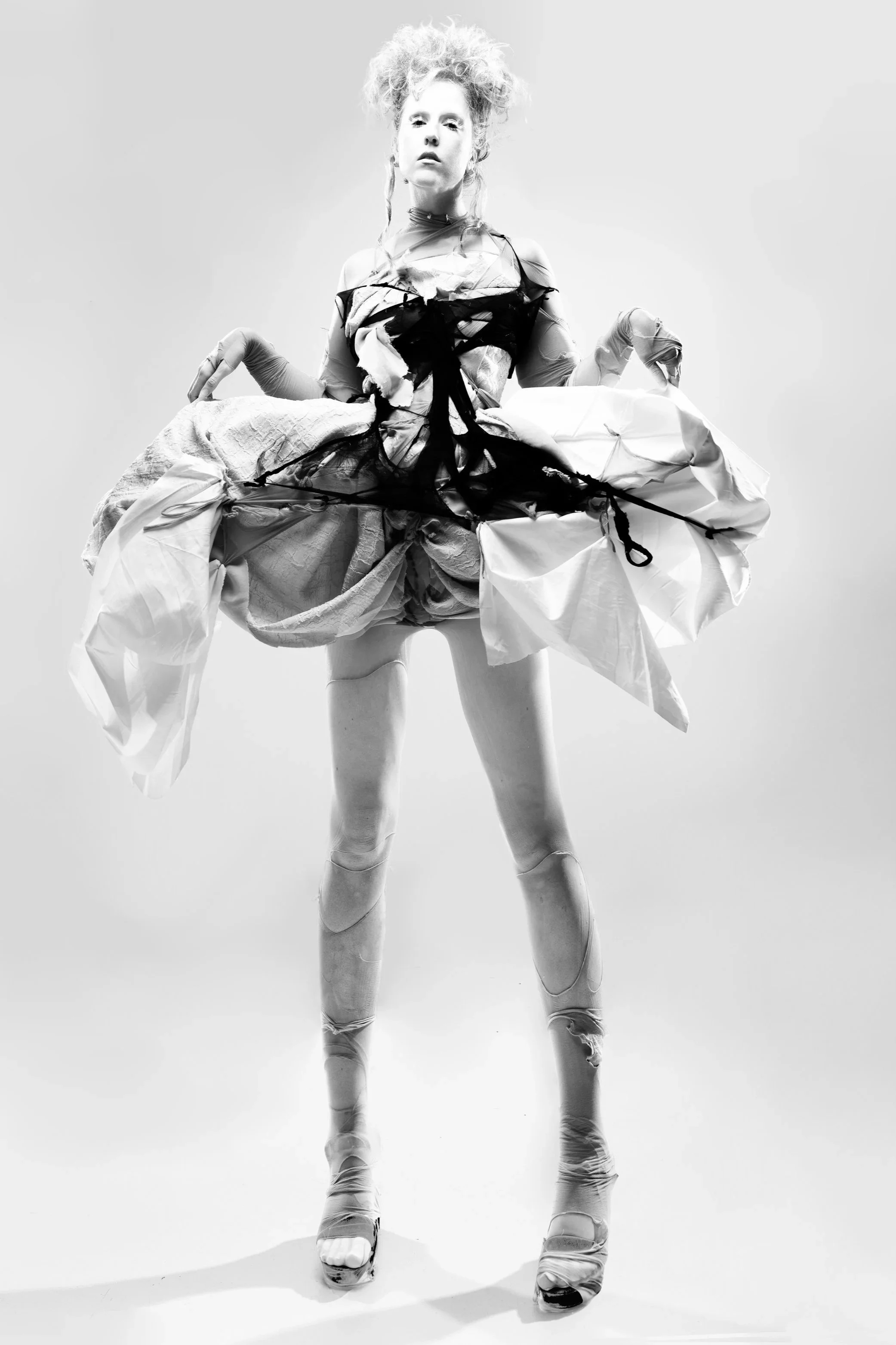 A woman with curly hair in avant-garde fashion poses in a studio with a plain background.