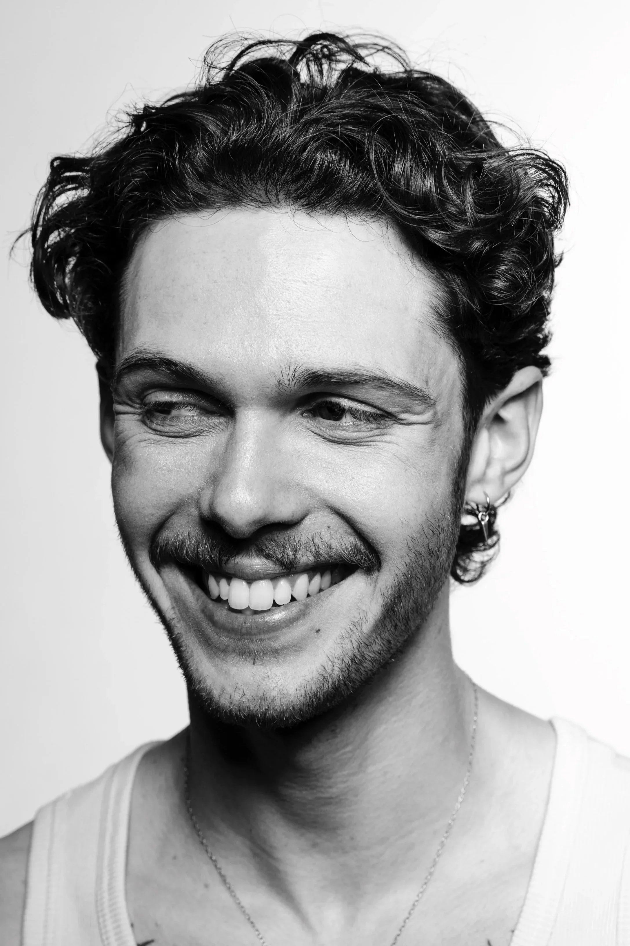 Black and white photo of a man with curly hair, a beard, and an earring, smiling with one eye closed.