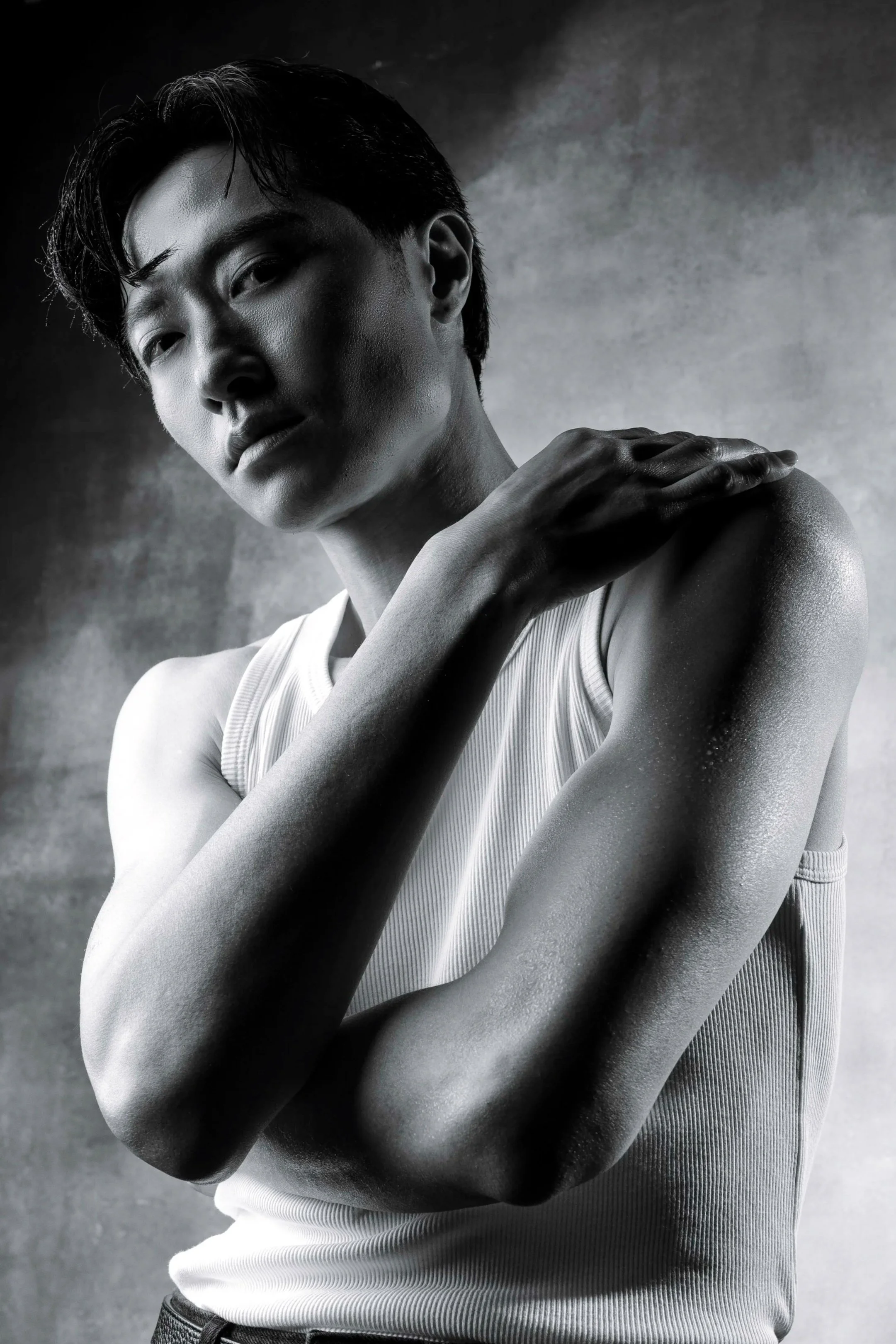 Black and white portrait of a young man with short, dark hair, wearing a sleeveless ribbed shirt and dark jeans, with one arm crossed over his shoulder, set against a plain textured background.