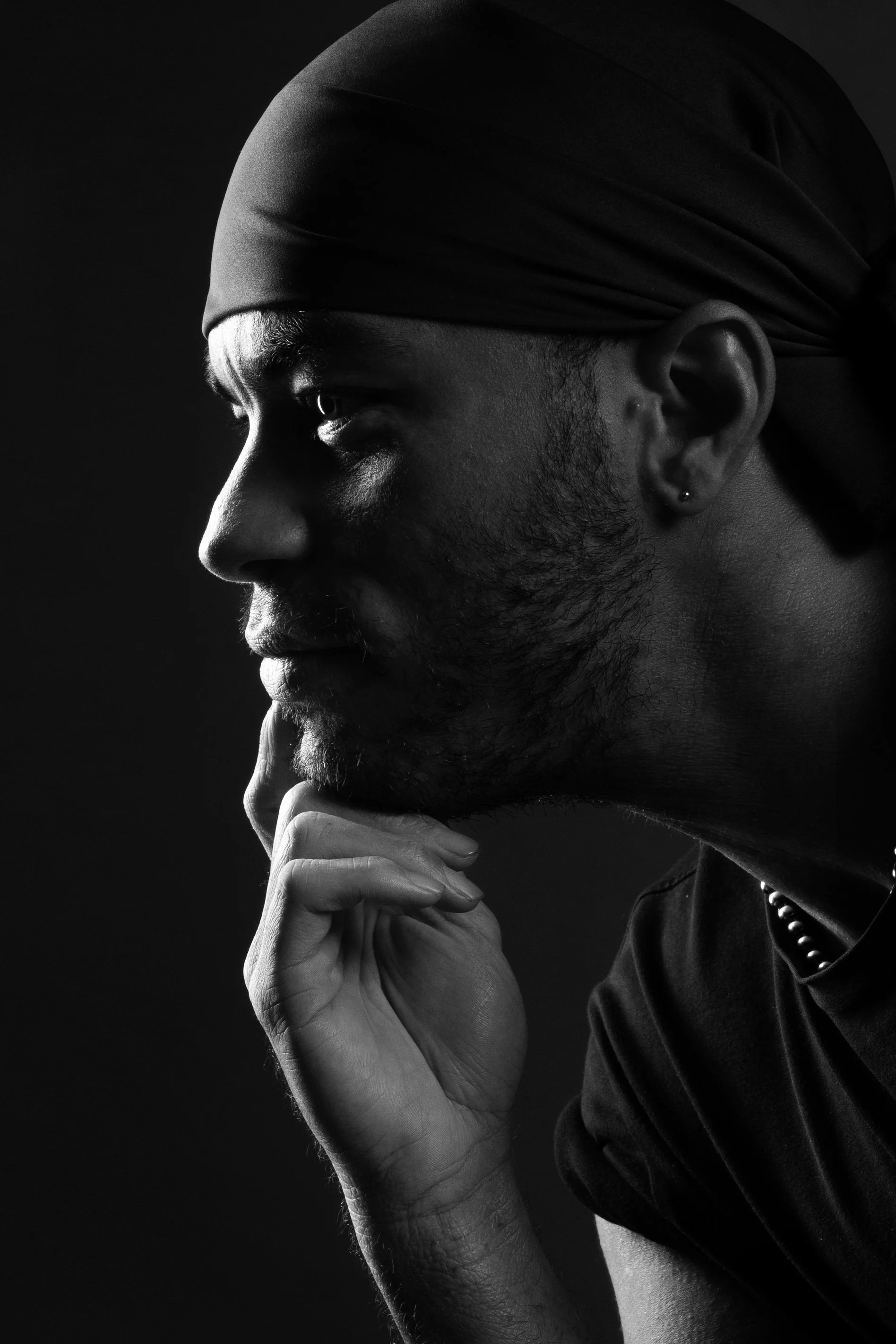 Black and white close-up portrait of a man wearing a headscarf, with thoughtful expression, hand resting under chin, dark background.