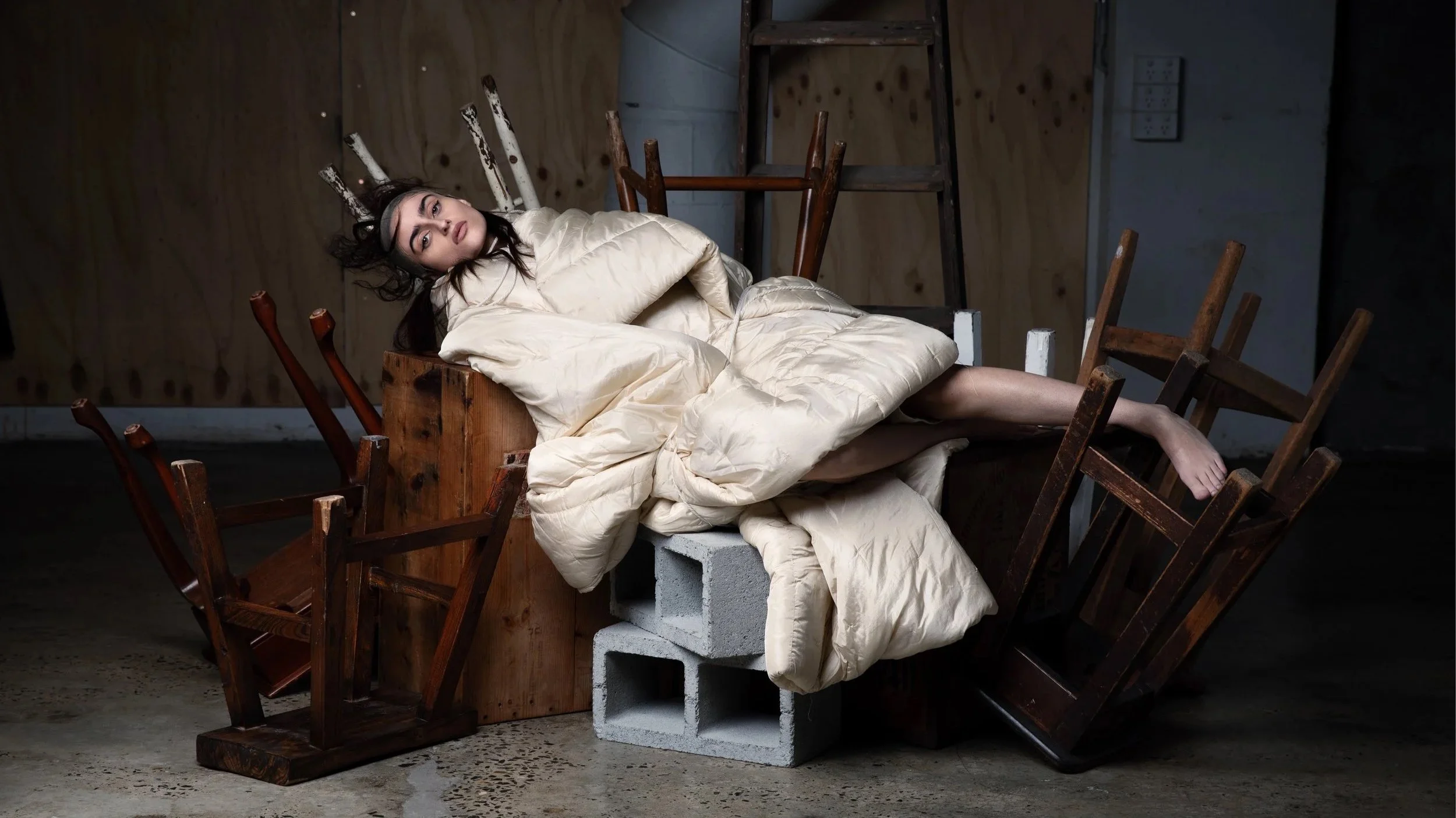 A woman lying on a pile of cream-colored bedding amid overturned wooden chairs and cinder blocks in a dimly lit, industrial-style room.