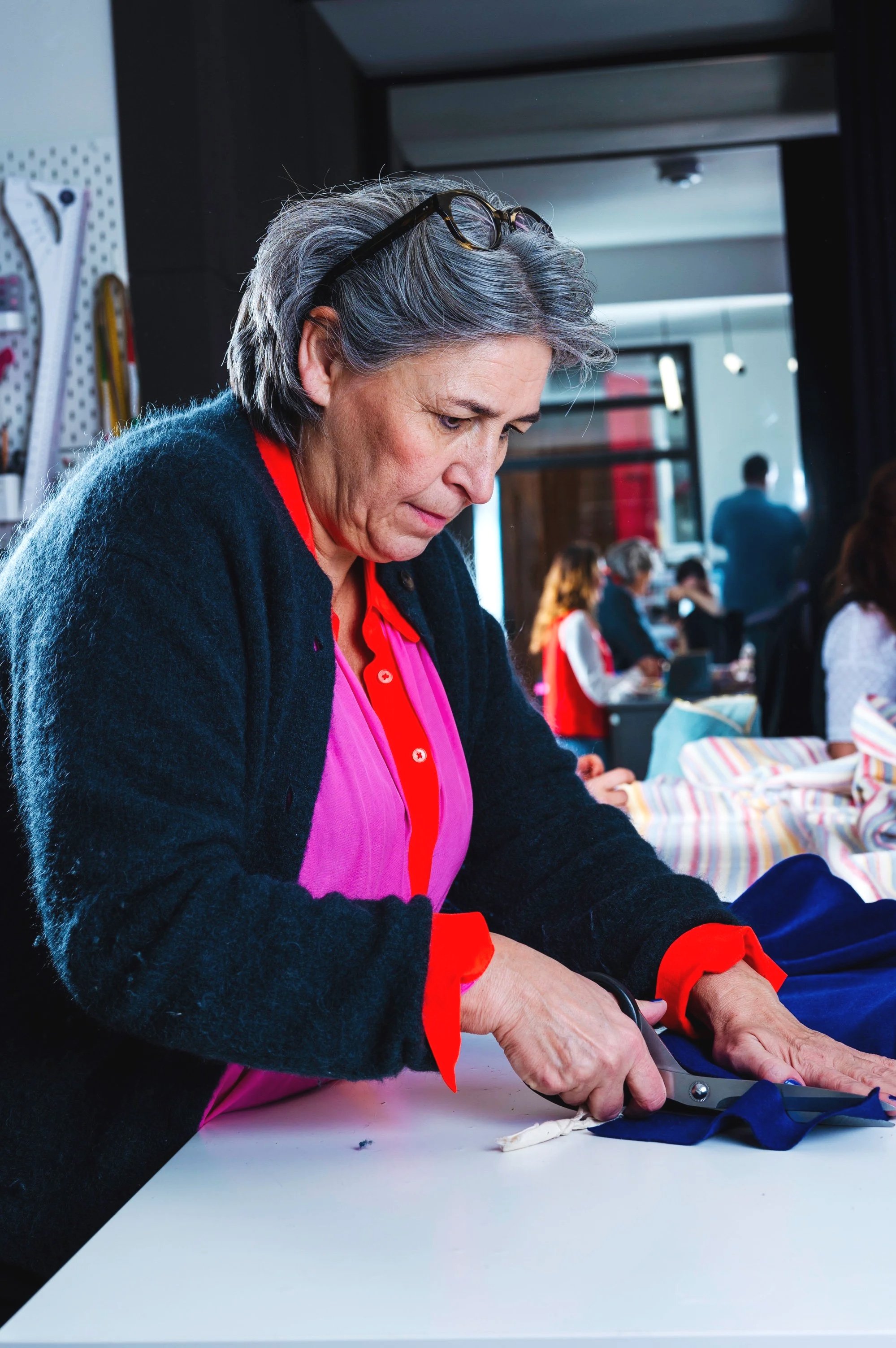 An older woman with glasses on her head is cutting fabric with scissors at a white table in a sewing workshop, with other people working in the background.