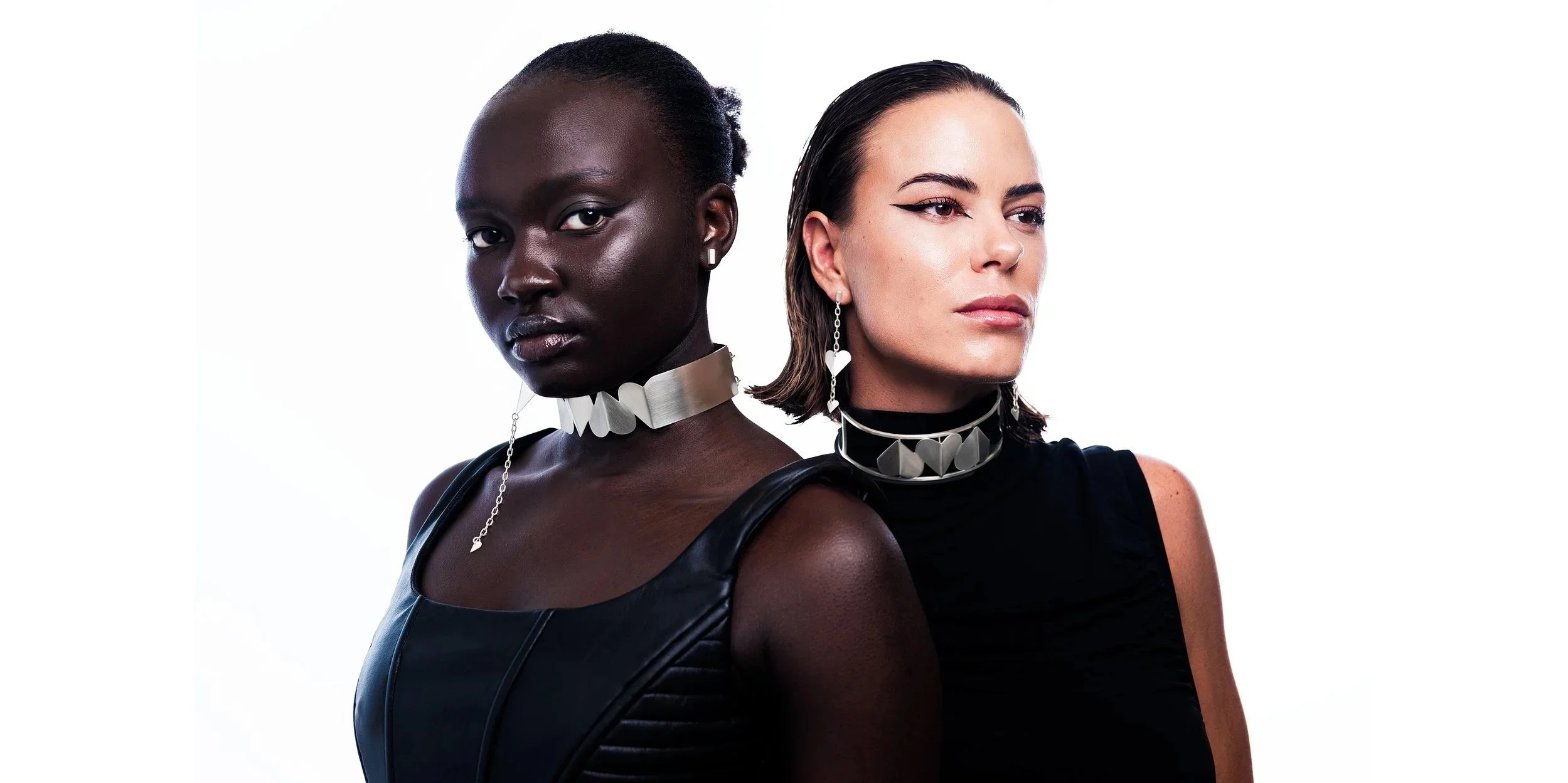 Two women wearing black outfits and matching metallic accessories, standing with serious expressions against a white background.