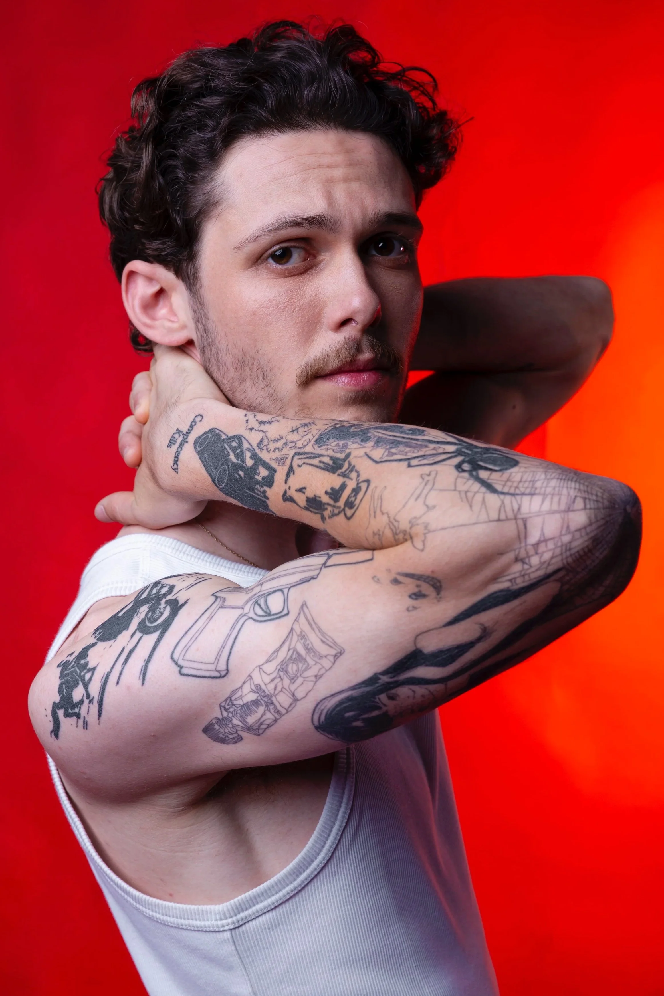 A young man with tattoos on his arms and chest, posing with one arm bent and his hand behind his head, against a red background.