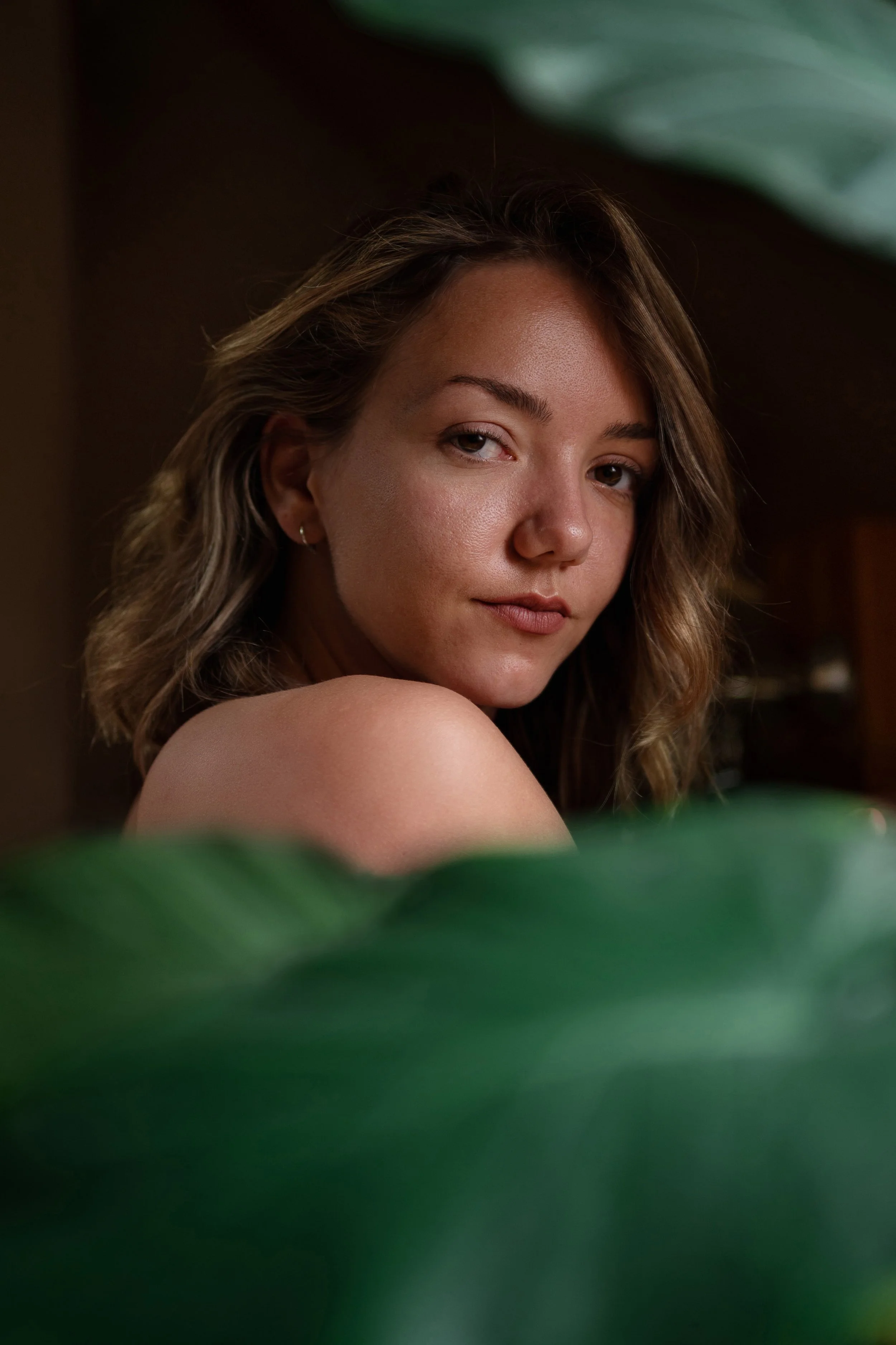 Close-up portrait of a young woman with wavy hair, looking at the camera with a slight smile, partially obscured by large green leaves in the foreground.