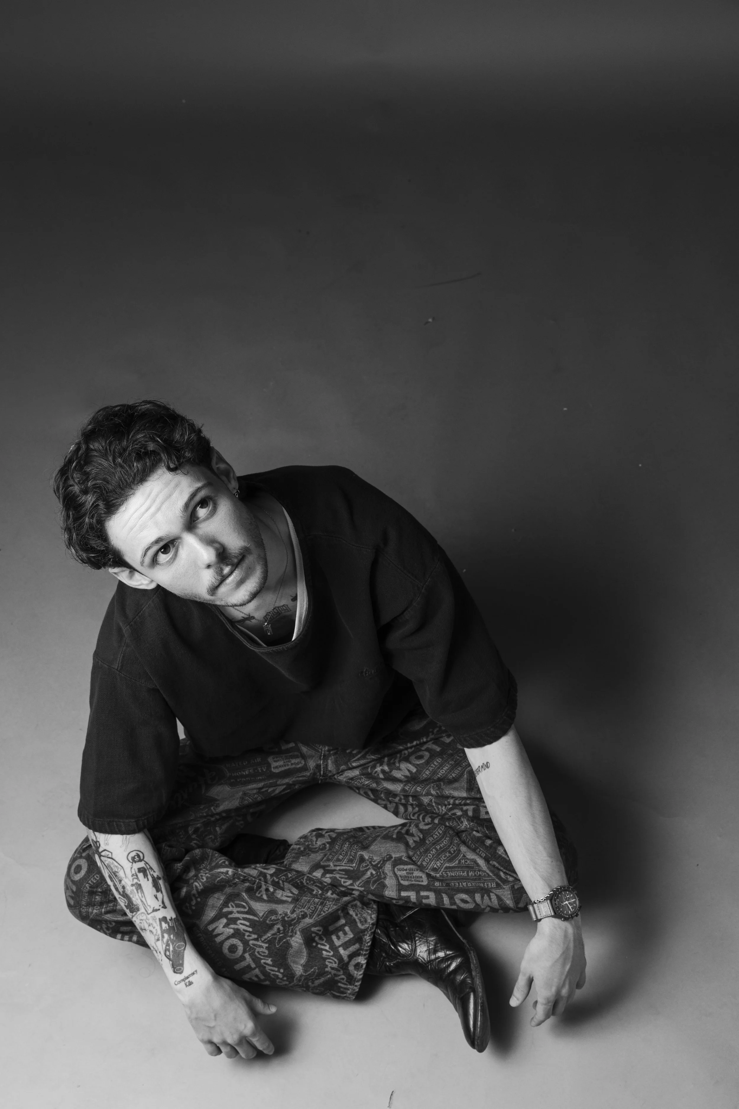 Black and white photo of a young man with tattoos, sitting cross-legged on the floor, looking up at the camera. He has curly hair, wearing a black shirt, patterned pants, and a watch.
