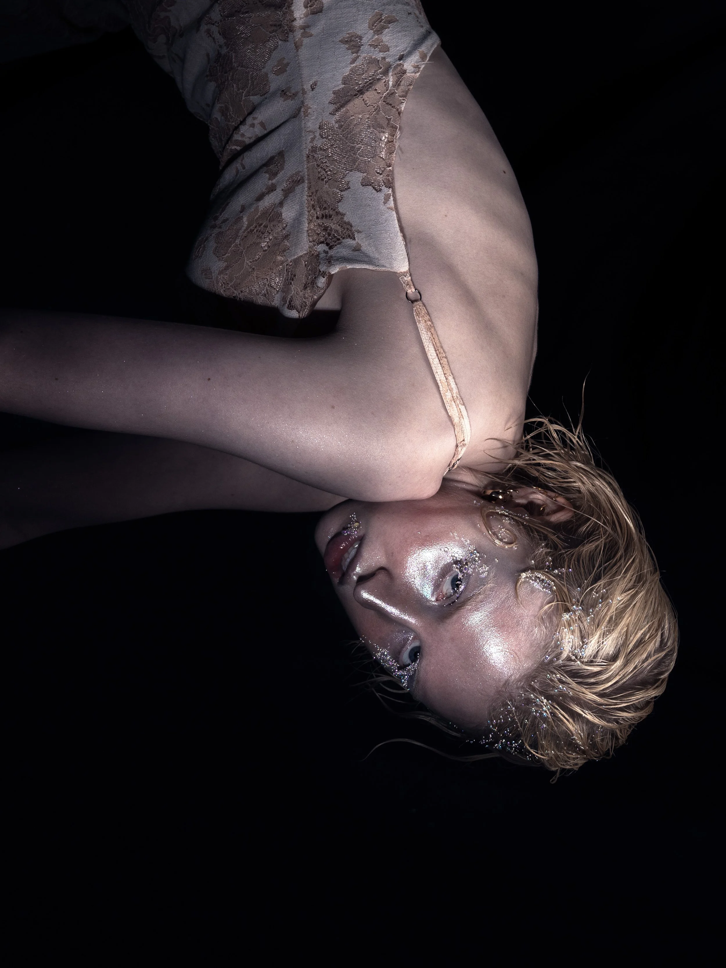 A woman with wet blonde hair lying on her side against a black background, wearing a sheer, lacy top with delicate straps and covered in shimmering makeup and glitter.