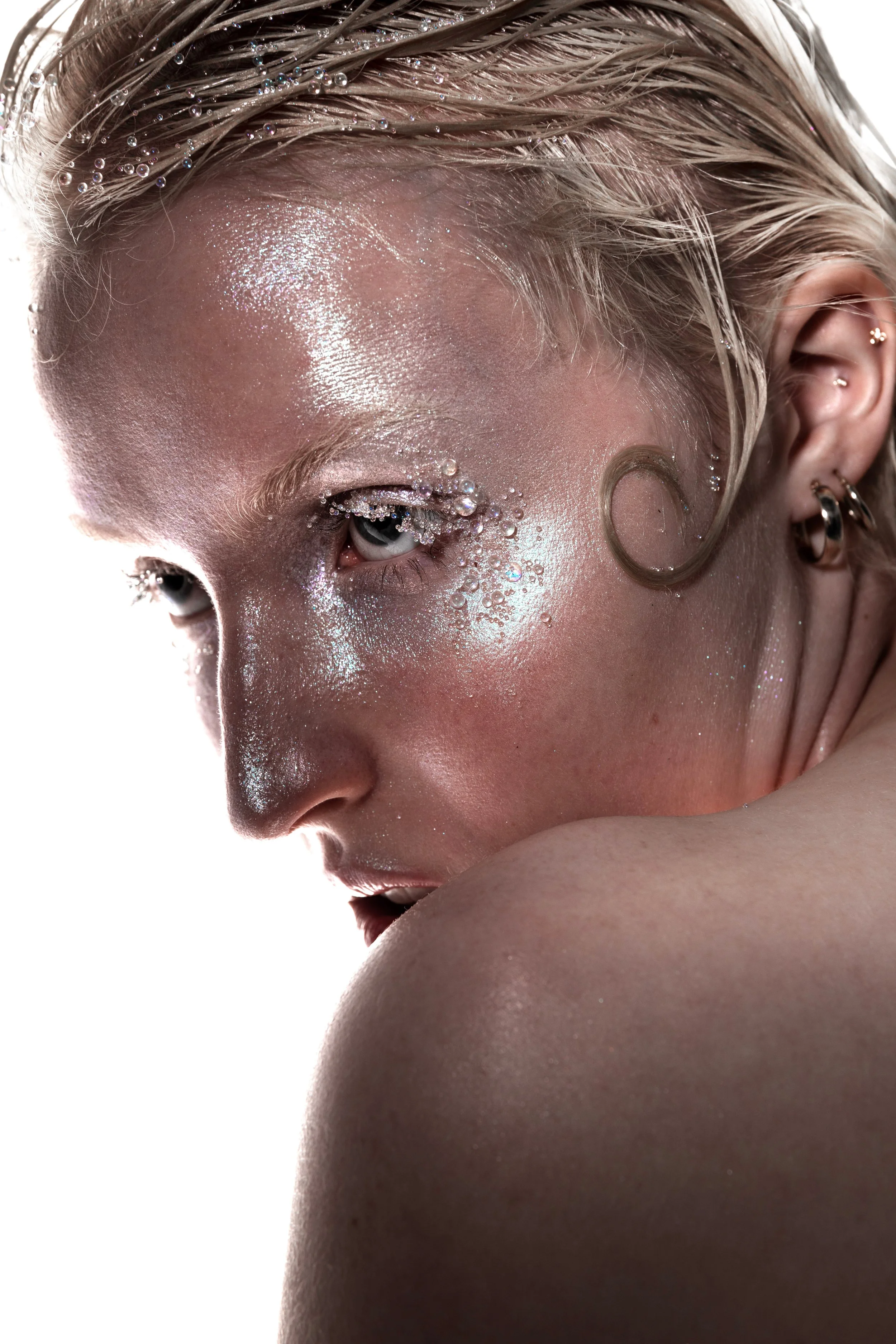 Close-up of a person with platinum blonde hair, glittery makeup, and pearl embellishments around the eye, with their shoulder visible.