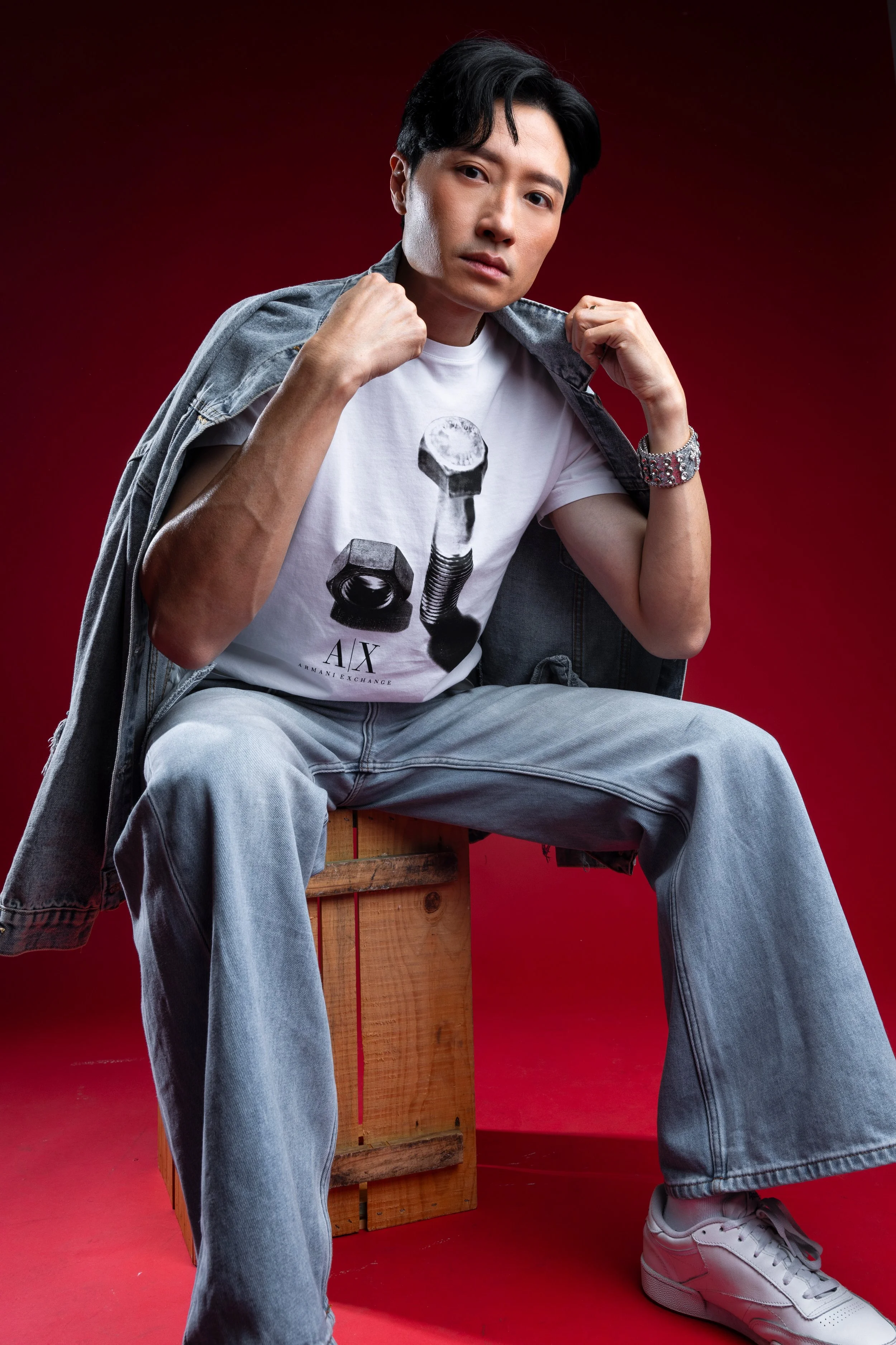 A young man wearing a white t-shirt with a graphic of a bolt and nut, light wash jeans, white sneakers, and a gray denim jacket draped over his shoulders, sitting on a wooden crate against a red background, looking at the camera.