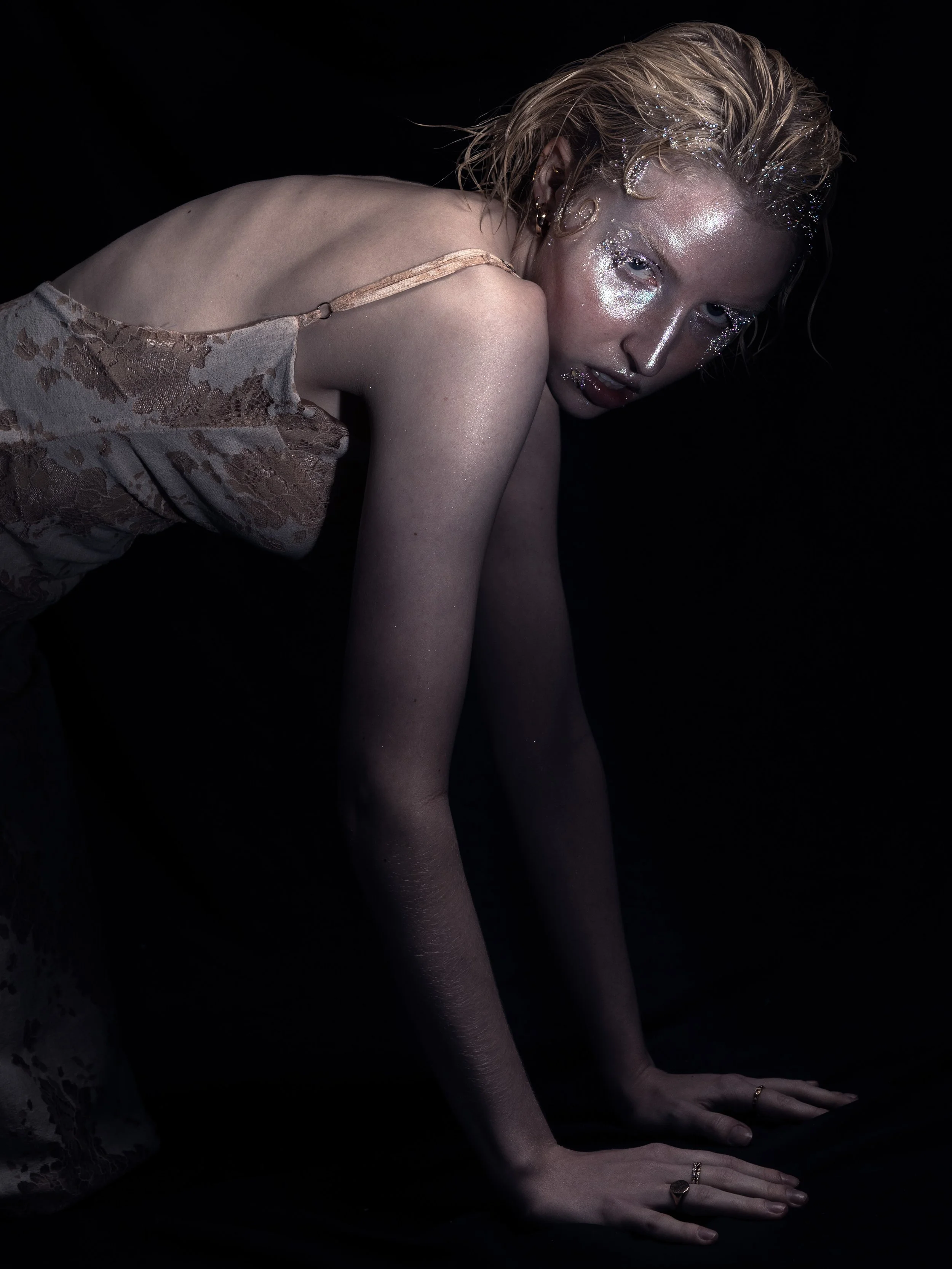 A person with short, wet, blond hair with glitter and makeup, posed on their hands and knees against a black background, wearing a lace slip dress.