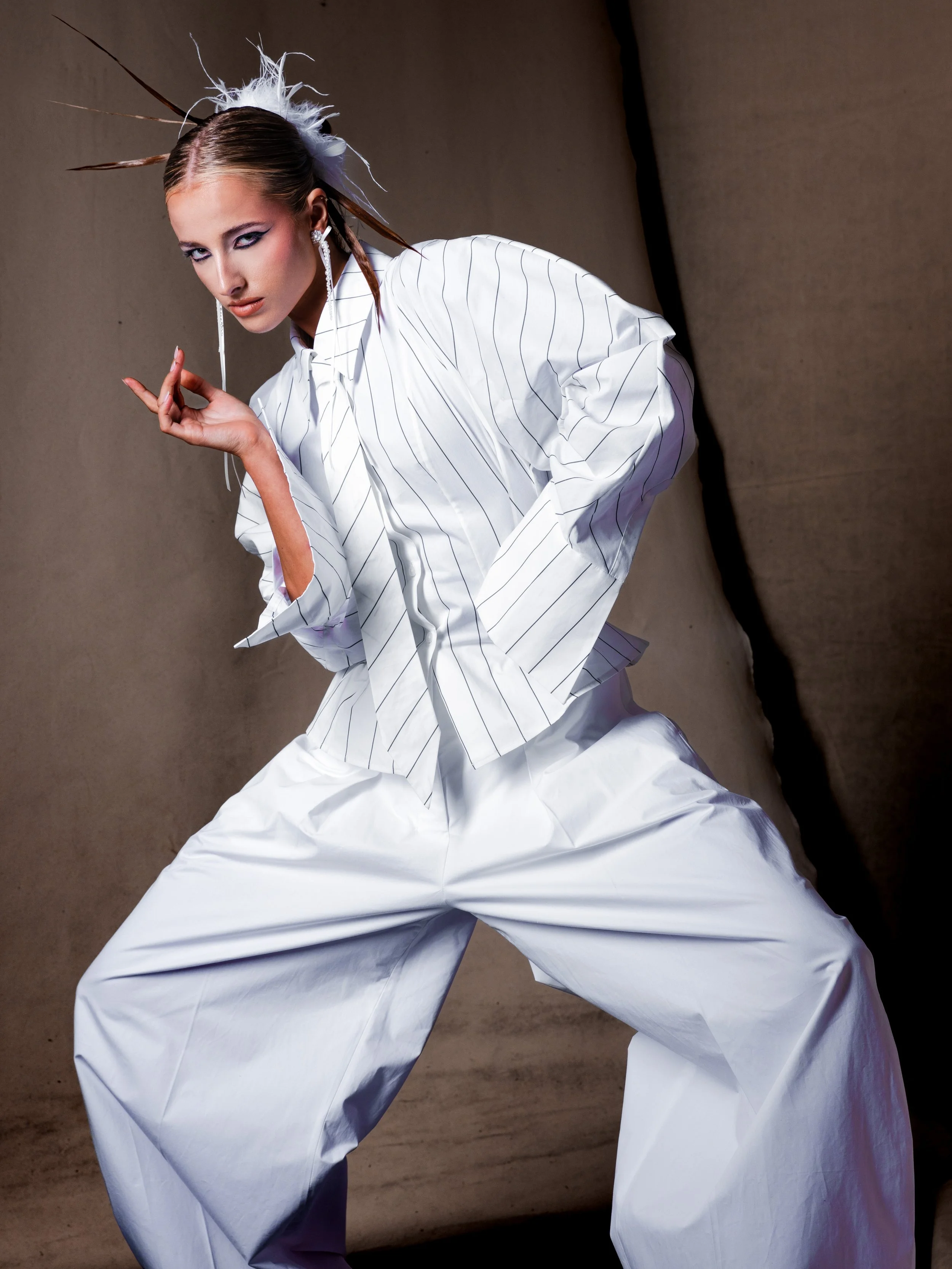 Fashion model dressed in white with pinstripe blazer, wide-leg pants, accessorized with feathers in hair, large earrings, striking makeup, posing against a plain background.