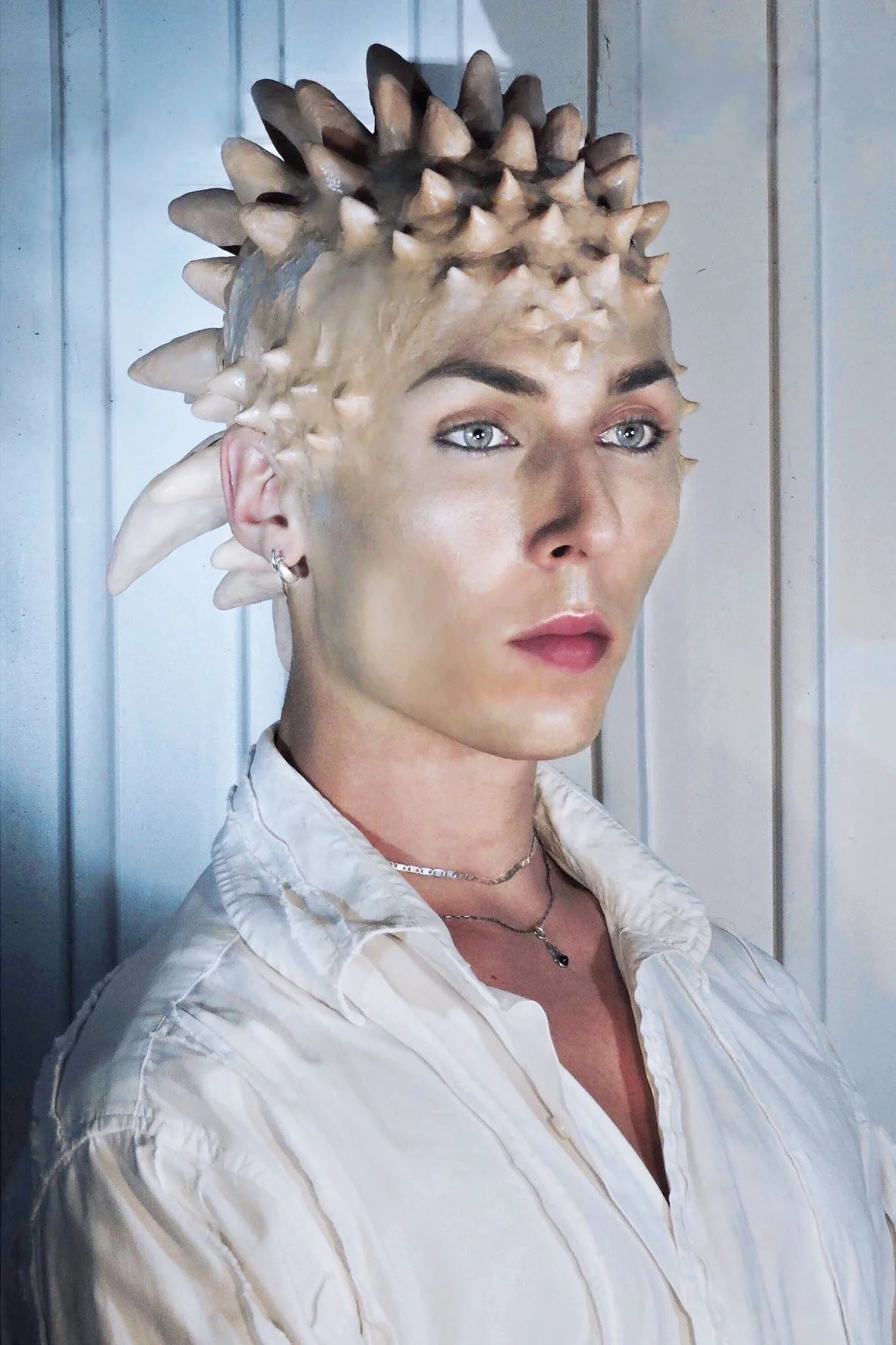Woman with Spiked Head Anemone-Style Hair wearing a white shirt and jewelry, standing in front of a wooden wall.