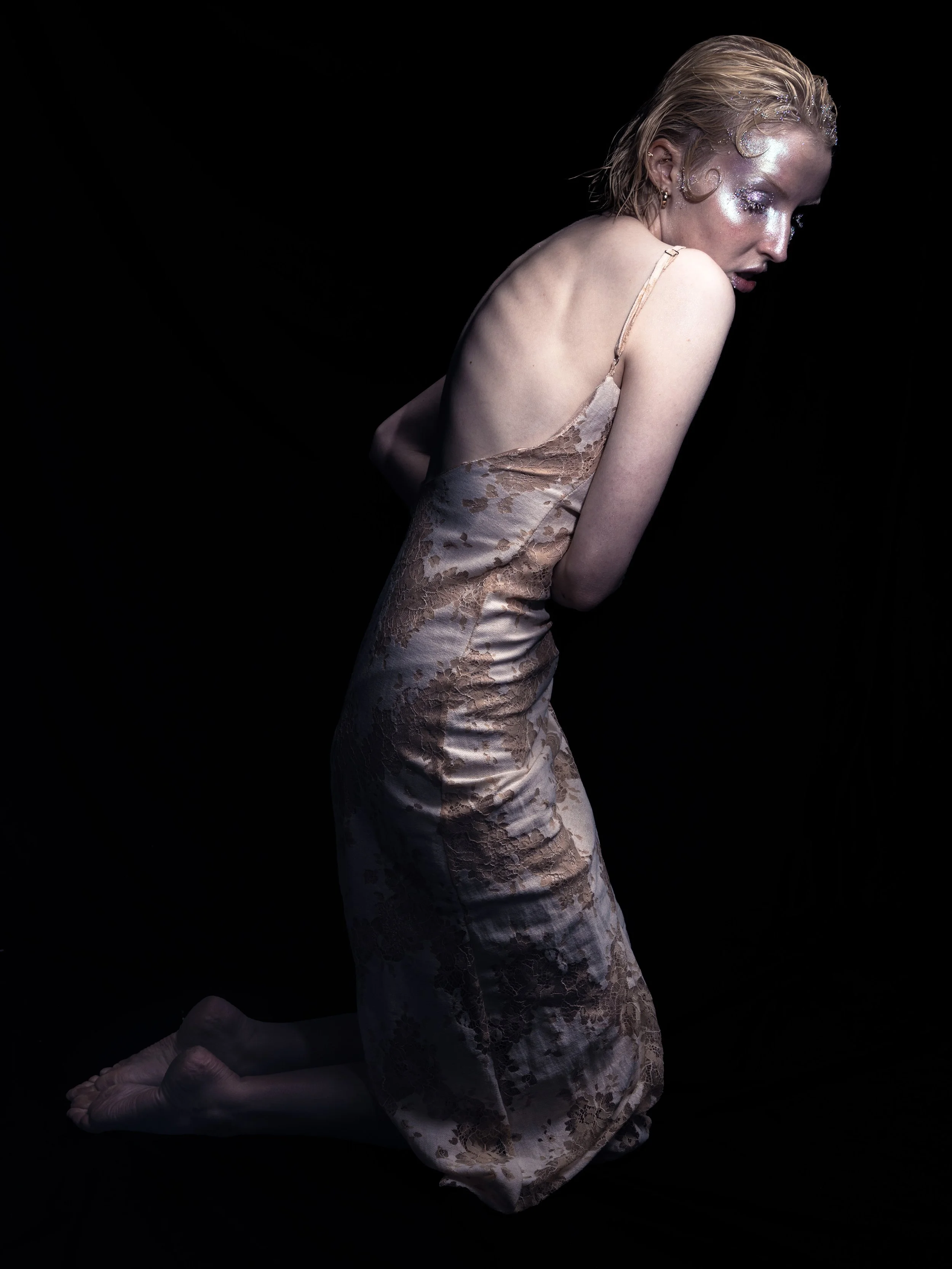 A woman with fair skin and short blonde hair kneeling on the floor against a black background, wearing a beige satin and lace slip dress, with makeup and glitter on her face.