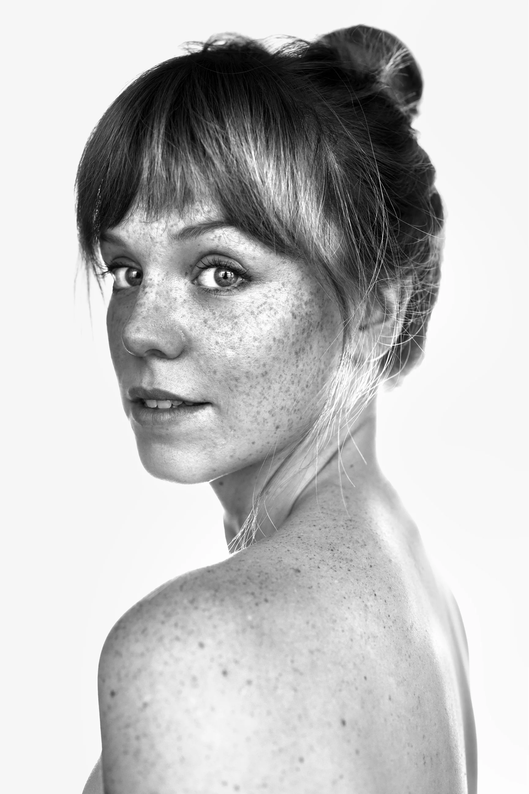 Close-up black and white portrait of a woman with freckles, looking over her shoulder with her hair in an updo.