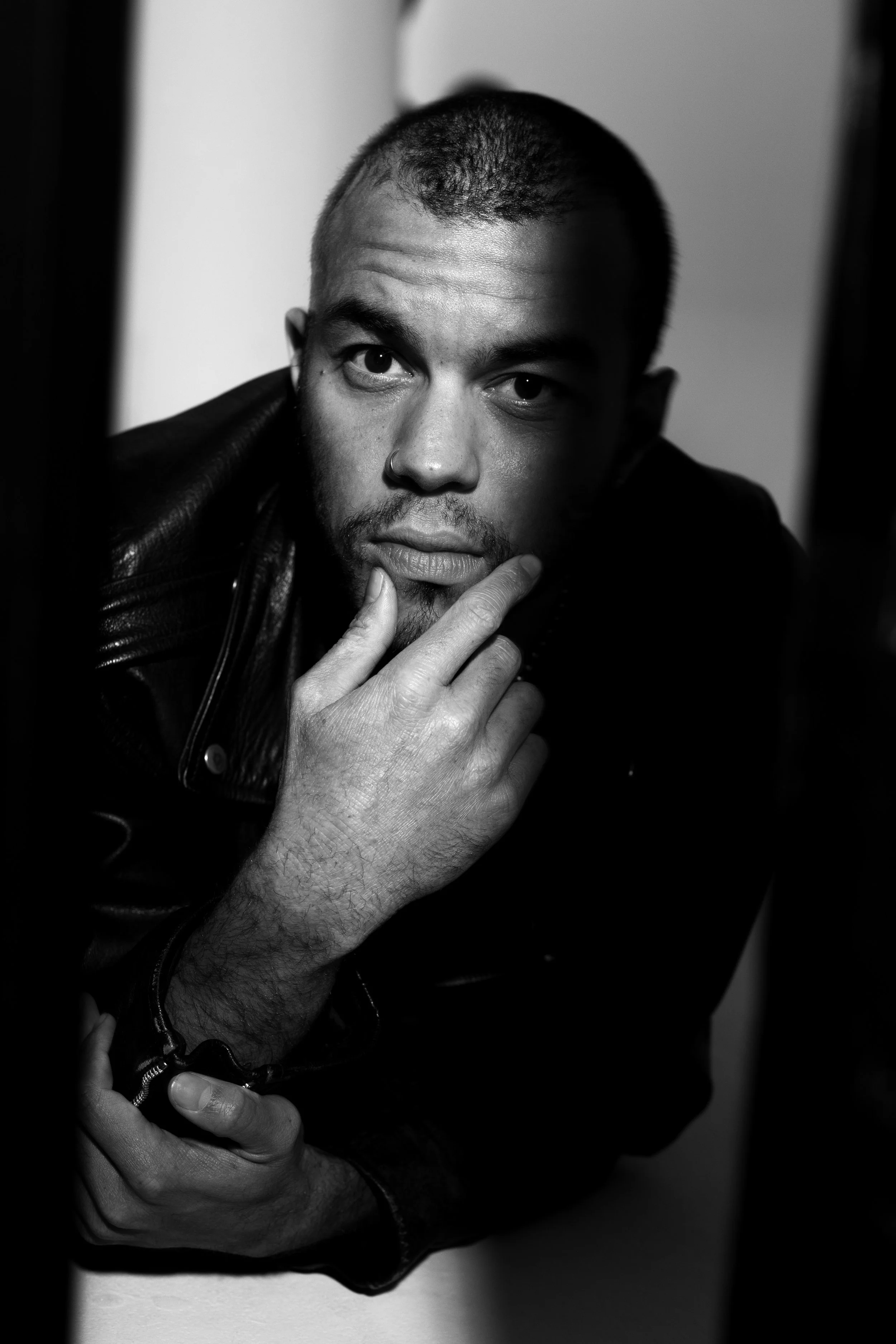 Black and white close-up portrait of a man with short hair, a nose ring, and a leather jacket, resting his chin on his hand and looking into the camera.