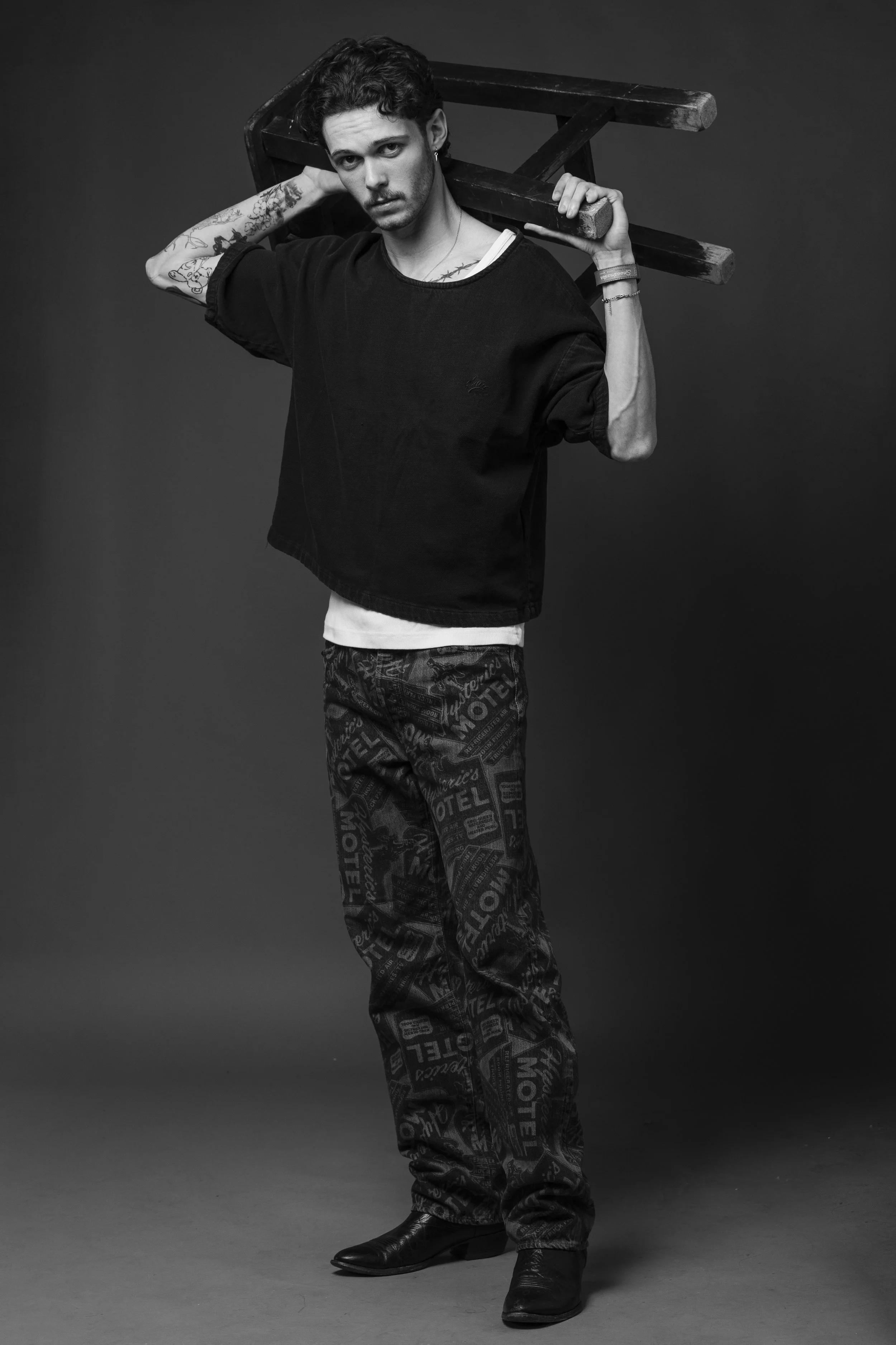 A young man with tattoos on his arms and a serious expression, holding a wooden chair over his shoulder, standing in a studio with a dark background, dressed in a black oversized shirt, patterned pants, and black boots.