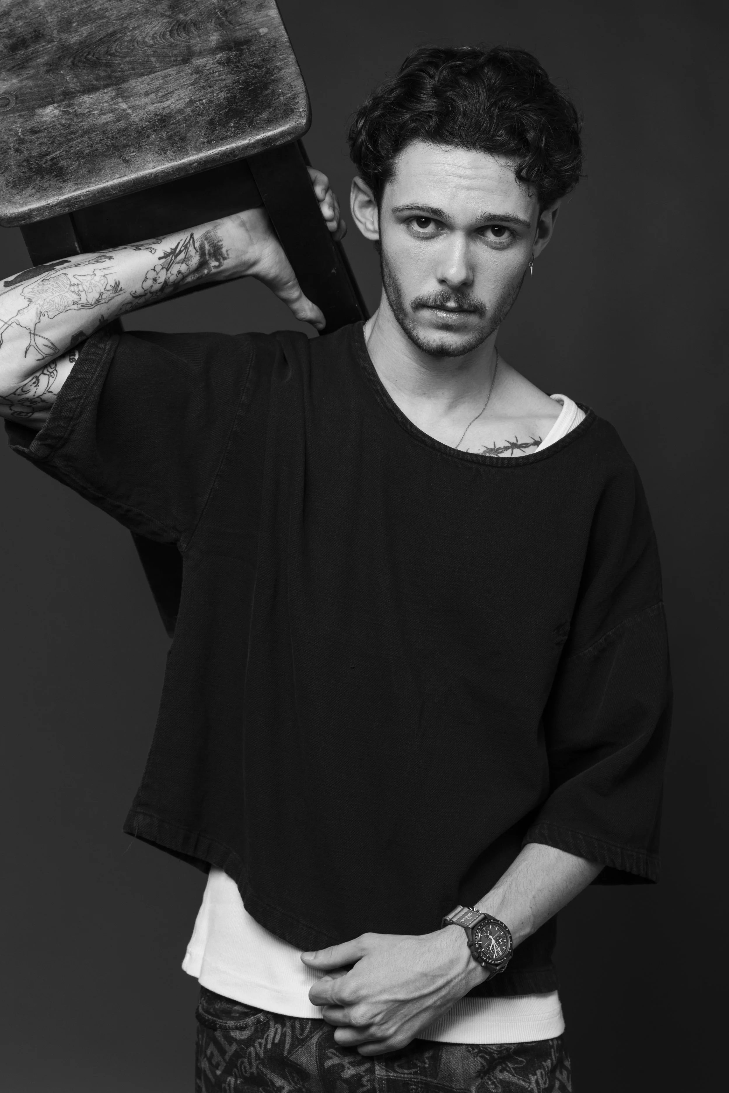 A black and white portrait of a young man with curly hair, tattoos on his arm, wearing a black oversized t-shirt, a wristwatch, and earrings, holding a wooden chair over his shoulder, against a plain background.