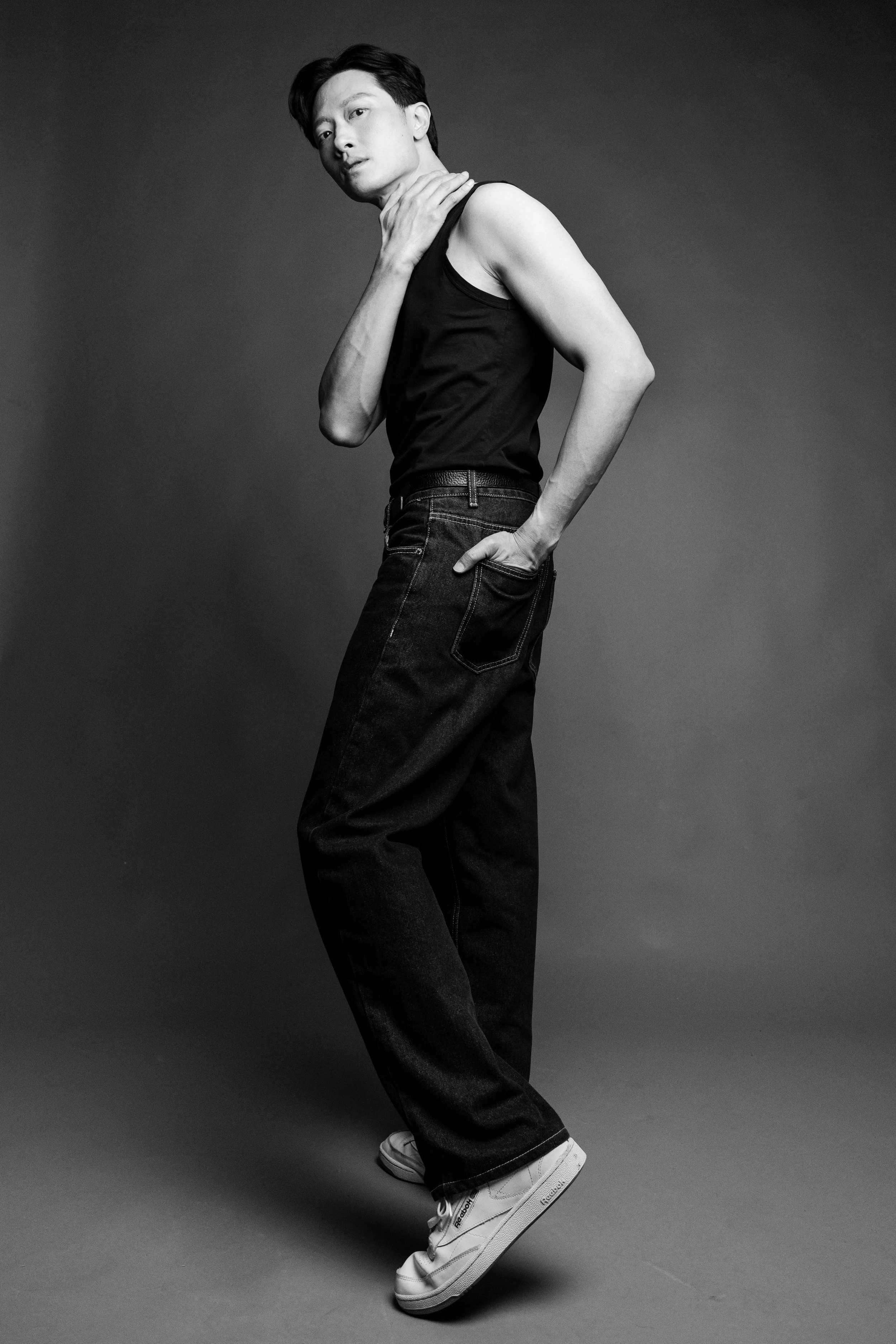 Black and white studio portrait of a young man posing with one hand on his neck and the other in his pocket, wearing a sleeveless top, jeans, and sneakers.