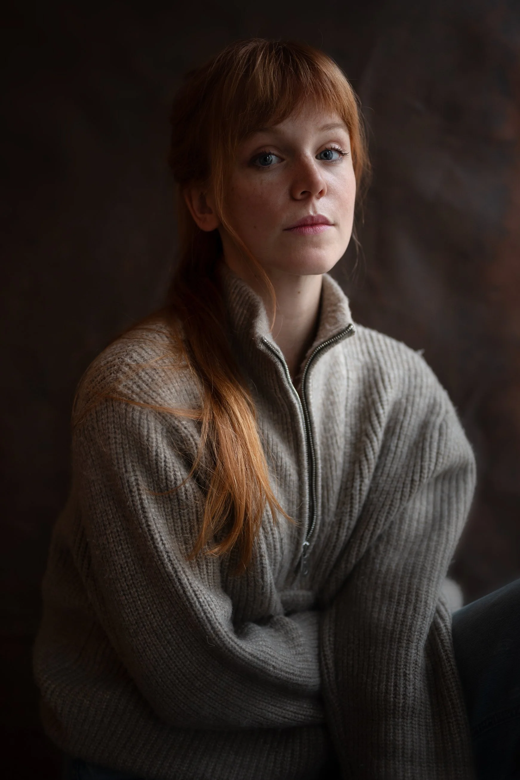 Portrait of a young woman with red hair wearing a beige zip-up sweater, sitting against a dark background.
