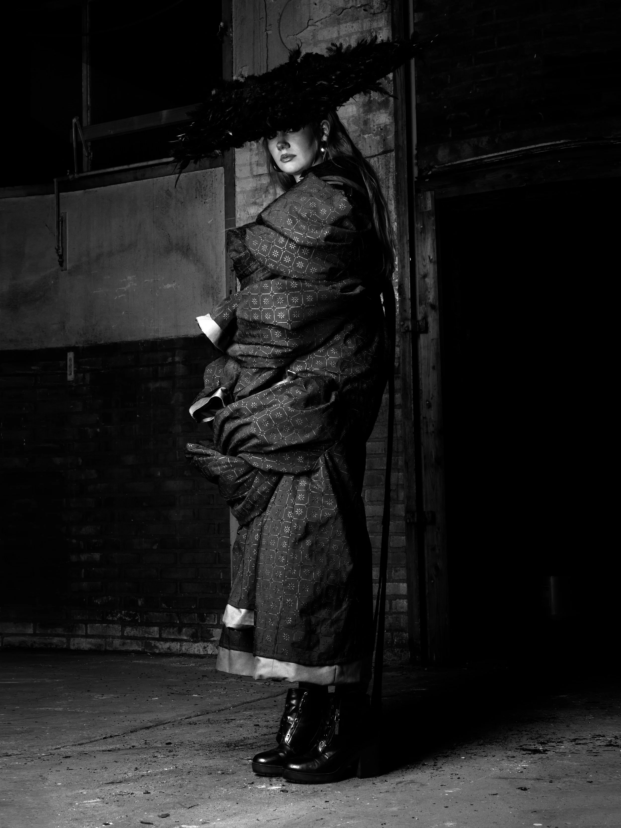 A woman dressed in fashionable clothing, standing against a brick wall in an industrial setting with a large feathered hat obscuring her eyes.