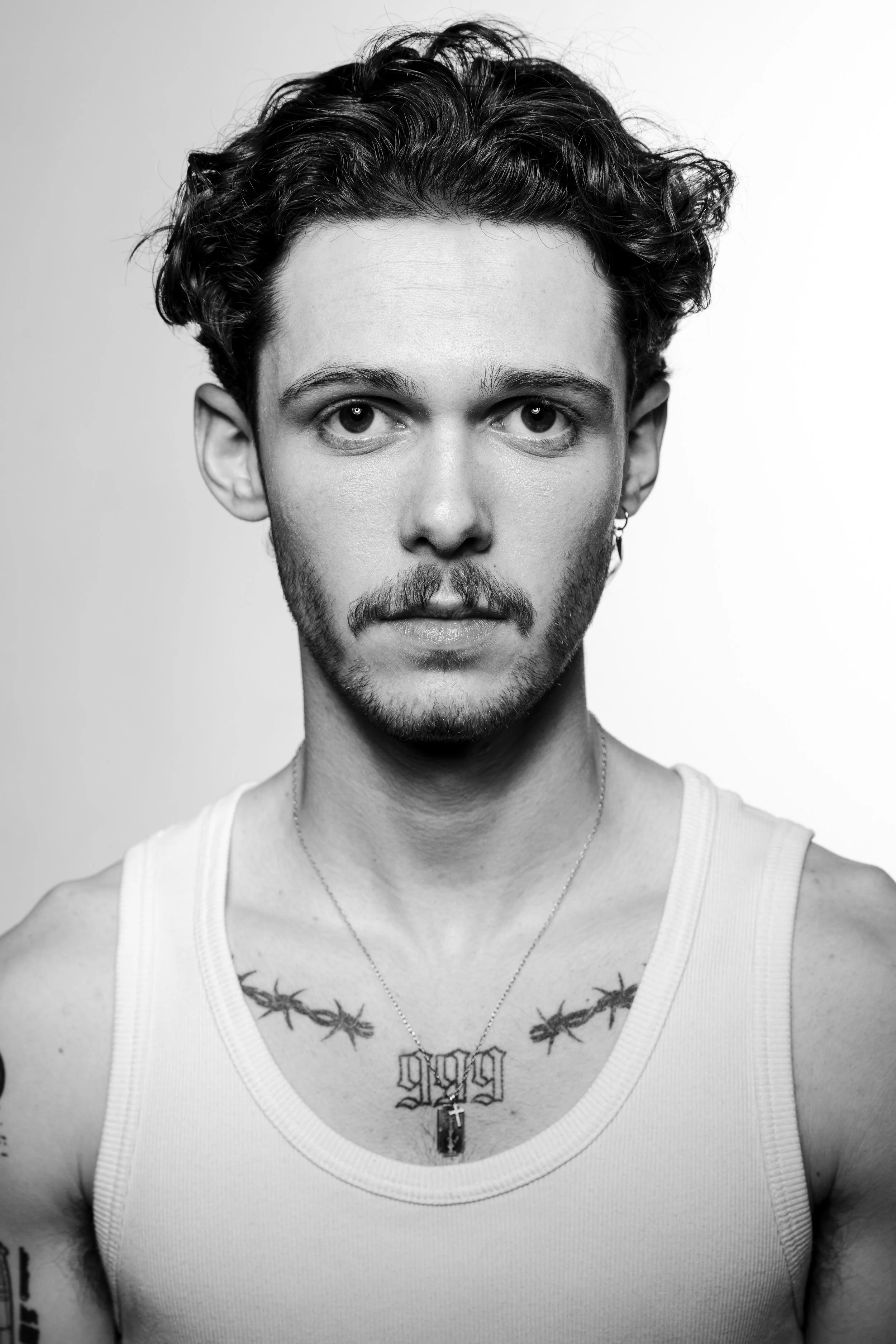Black and white portrait of a young man with short curly hair, stubble, wearing a white tank top, a necklace with a pendant, and earrings. He has tattoos on his chest and arms, and is looking directly at the camera with a neutral expression.