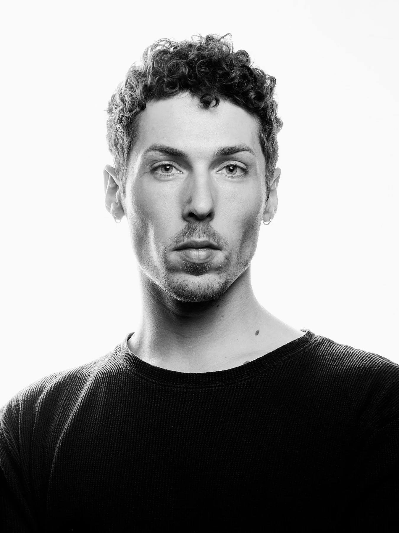 Black and white portrait of a man with short curly hair, light-colored eyes, wearing a dark textured shirt, looking directly at the camera.