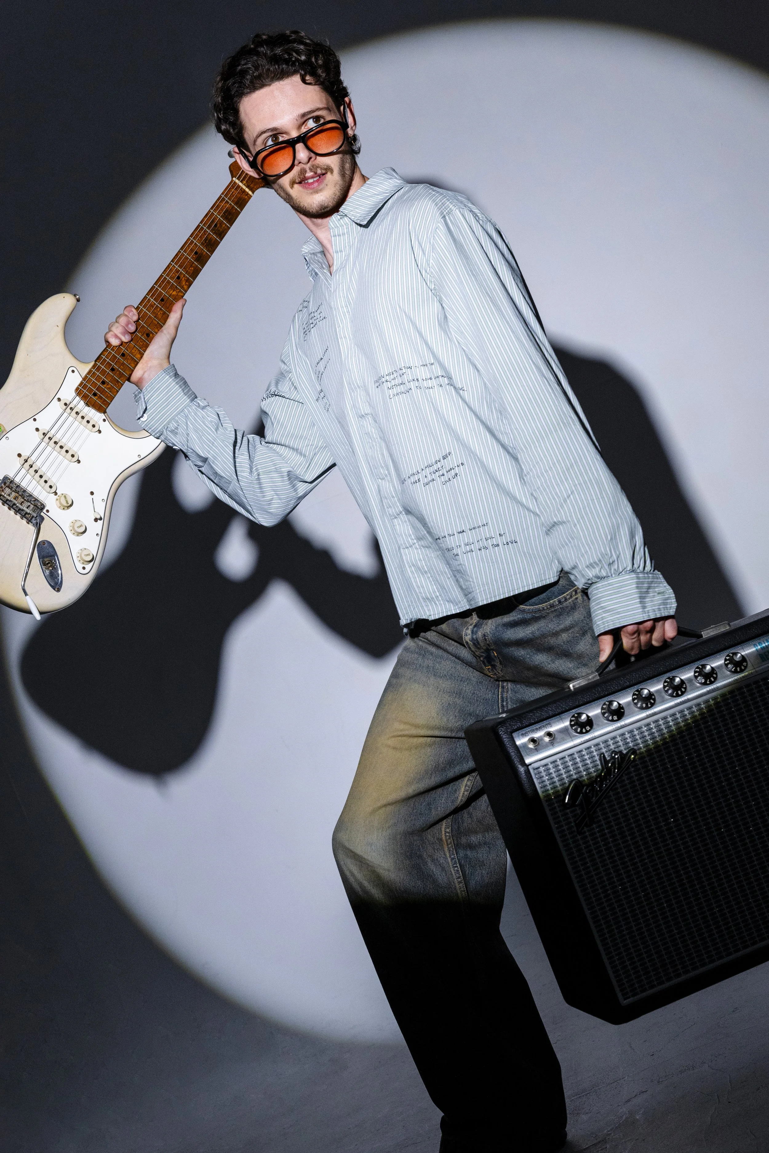 A young man with curly dark hair wearing orange-tinted glasses, a light striped button-up shirt, and jeans, holding a cream-colored electric guitar on his shoulder and a black guitar amplifier in his other hand, standing in front of a white circle pr