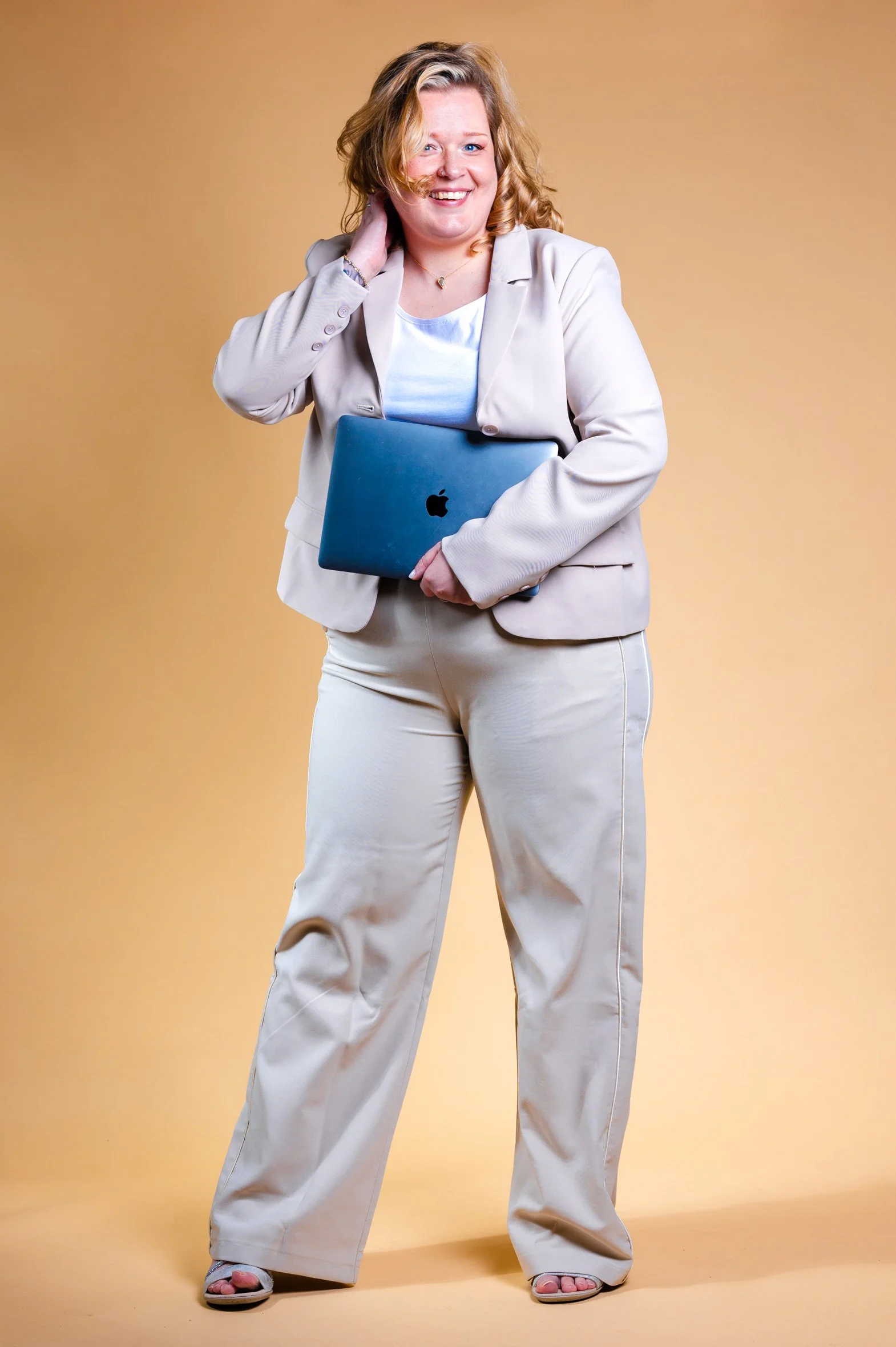 A woman with shoulder-length blonde hair smiling, holding a closed blue MacBook in her left hand and touching her neck with her right hand, standing against a plain beige background.