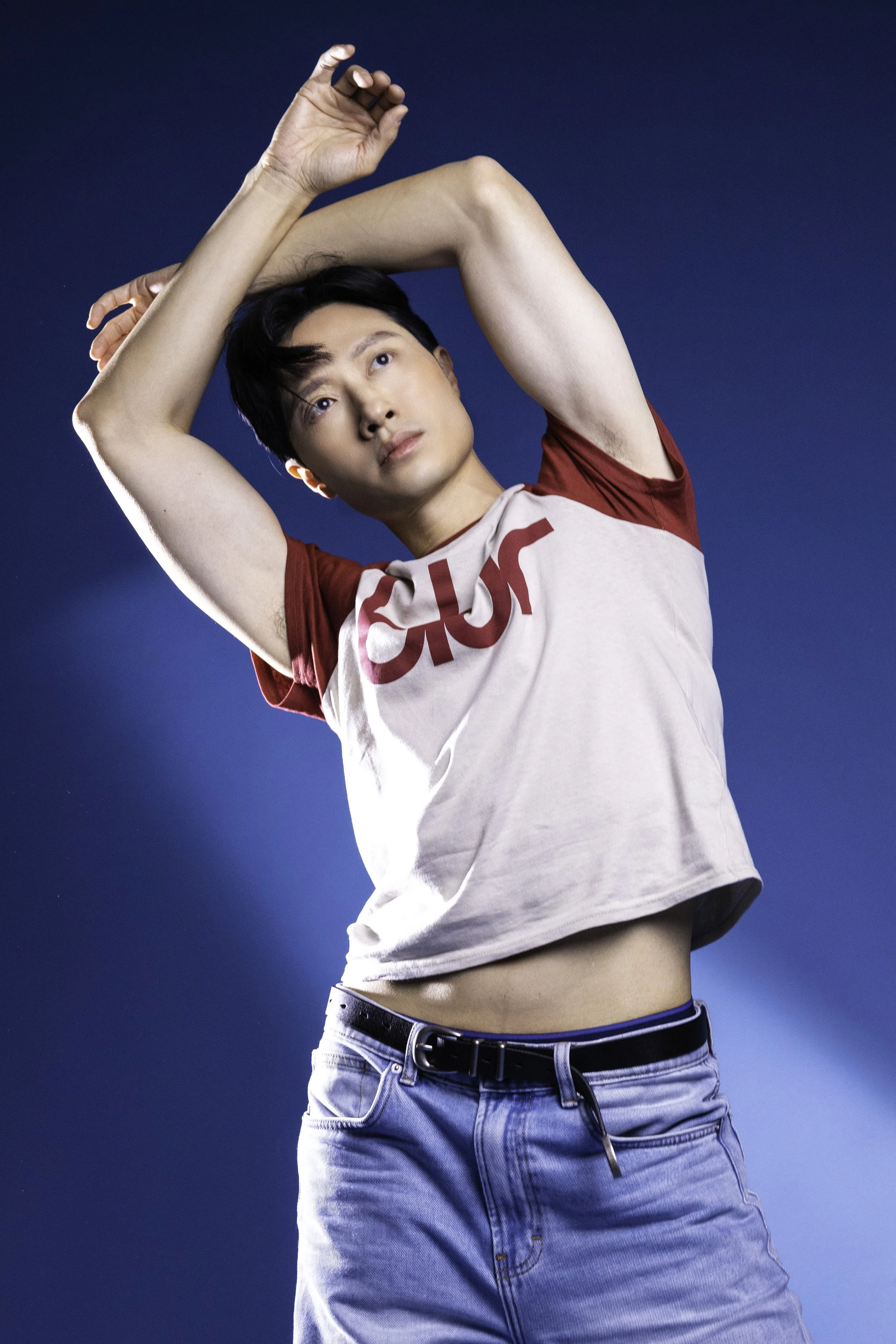 A young person with short dark hair, wearing a white and red t-shirt and blue jeans, striking a pose with arms above their head against a blue background.