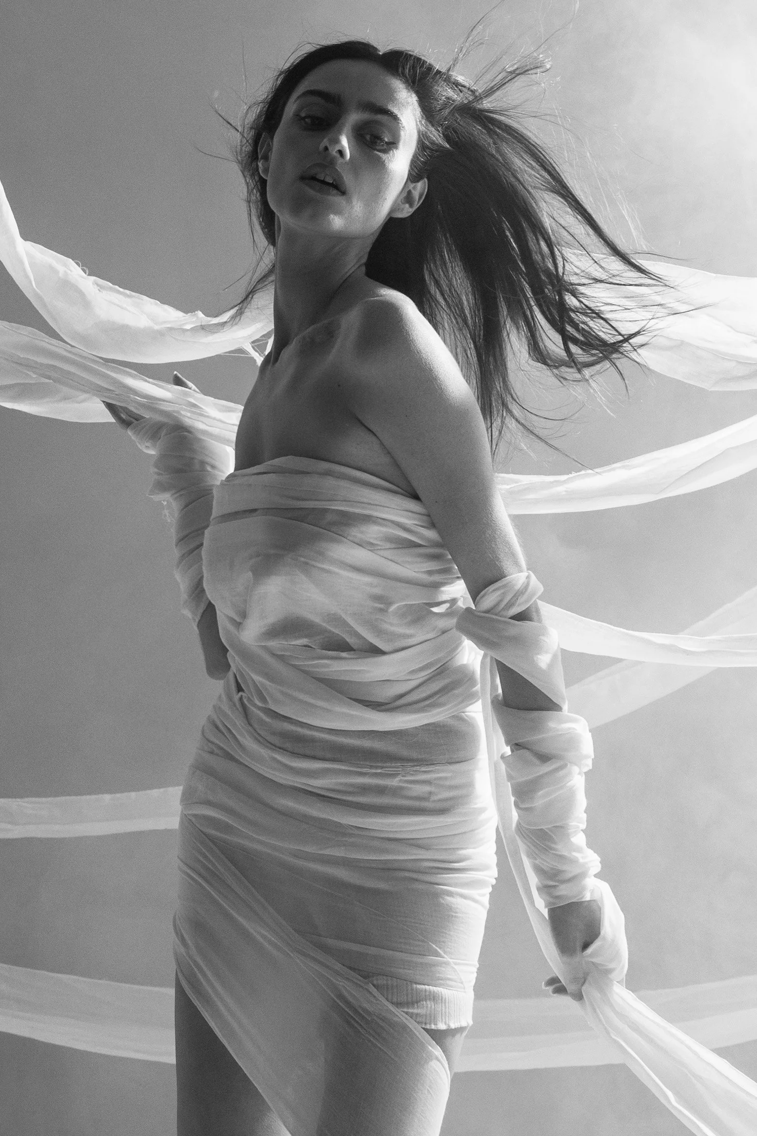 A woman with long hair blowing in the wind wrapped in flowing fabric, looking at the camera in a black and white photo.