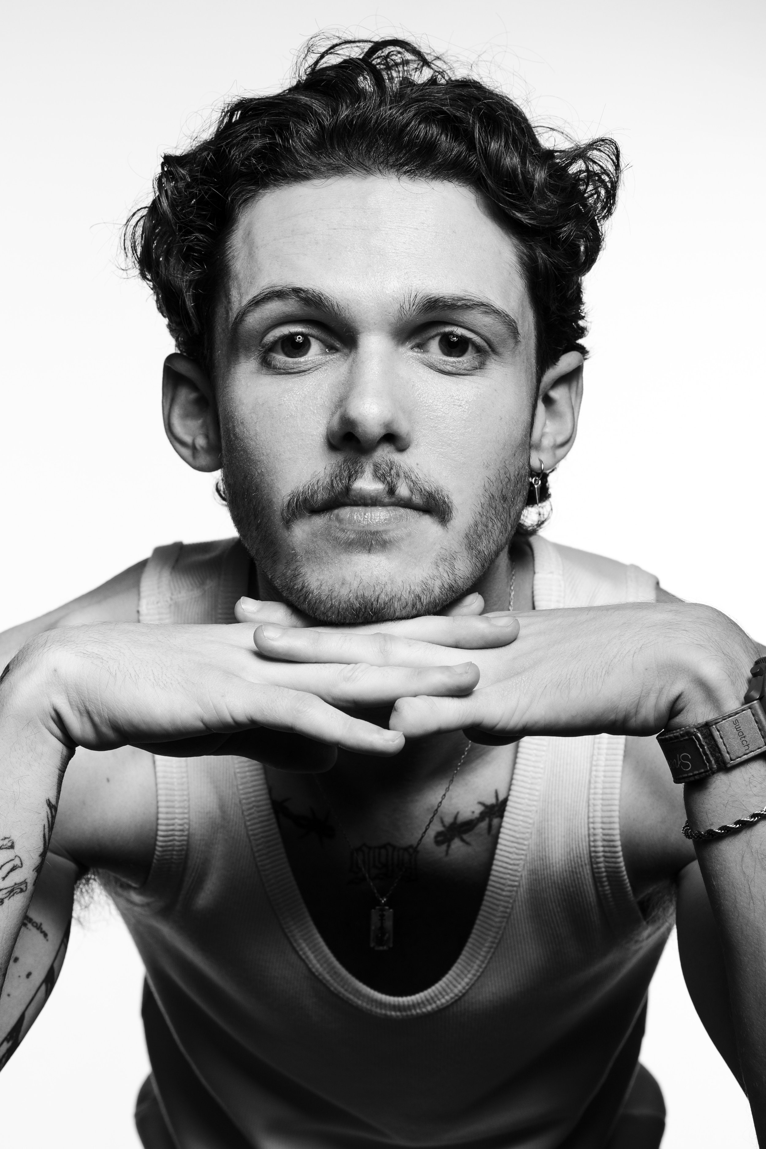 A black-and-white portrait of a young man with curly hair and a beard, resting his chin on his folded hands, wearing a sleeveless shirt, earrings, a watch, and a necklace.