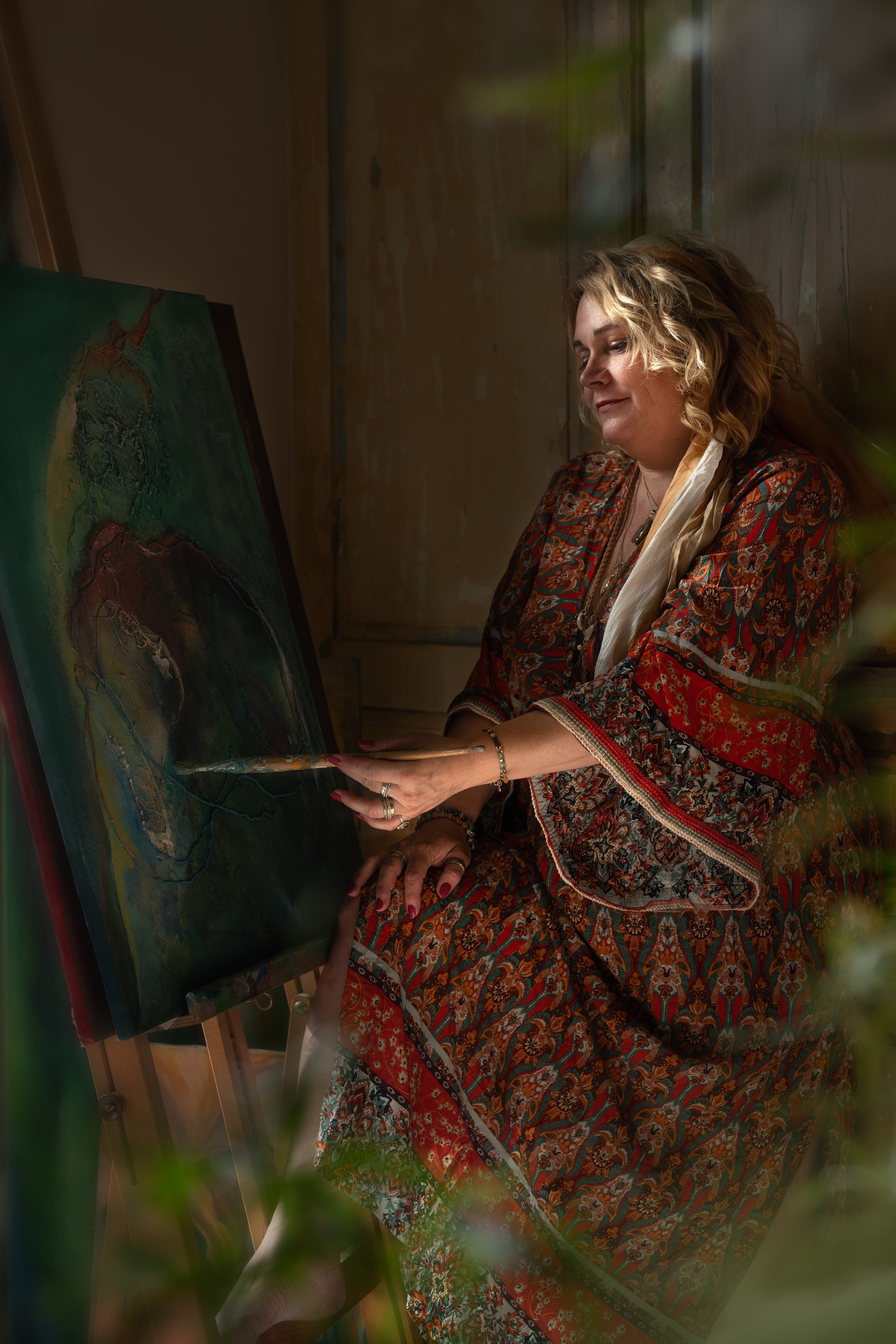A woman with curly blonde hair, wearing a patterned dress, is sitting in front of an abstract painting and working on it with a brush.