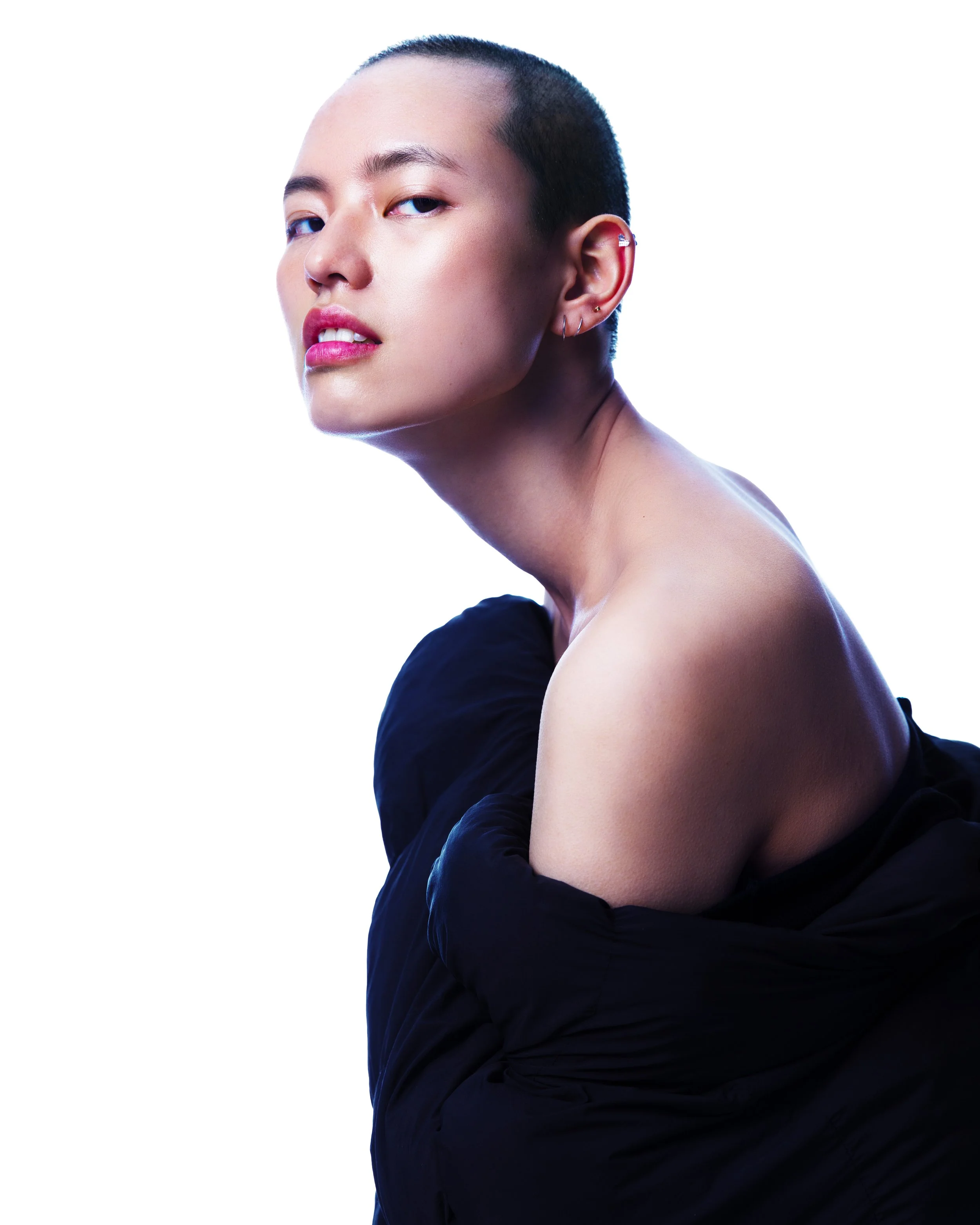 Portrait of a person with a shaved head, wearing small earrings, partially covered with a black garment, against a white background.