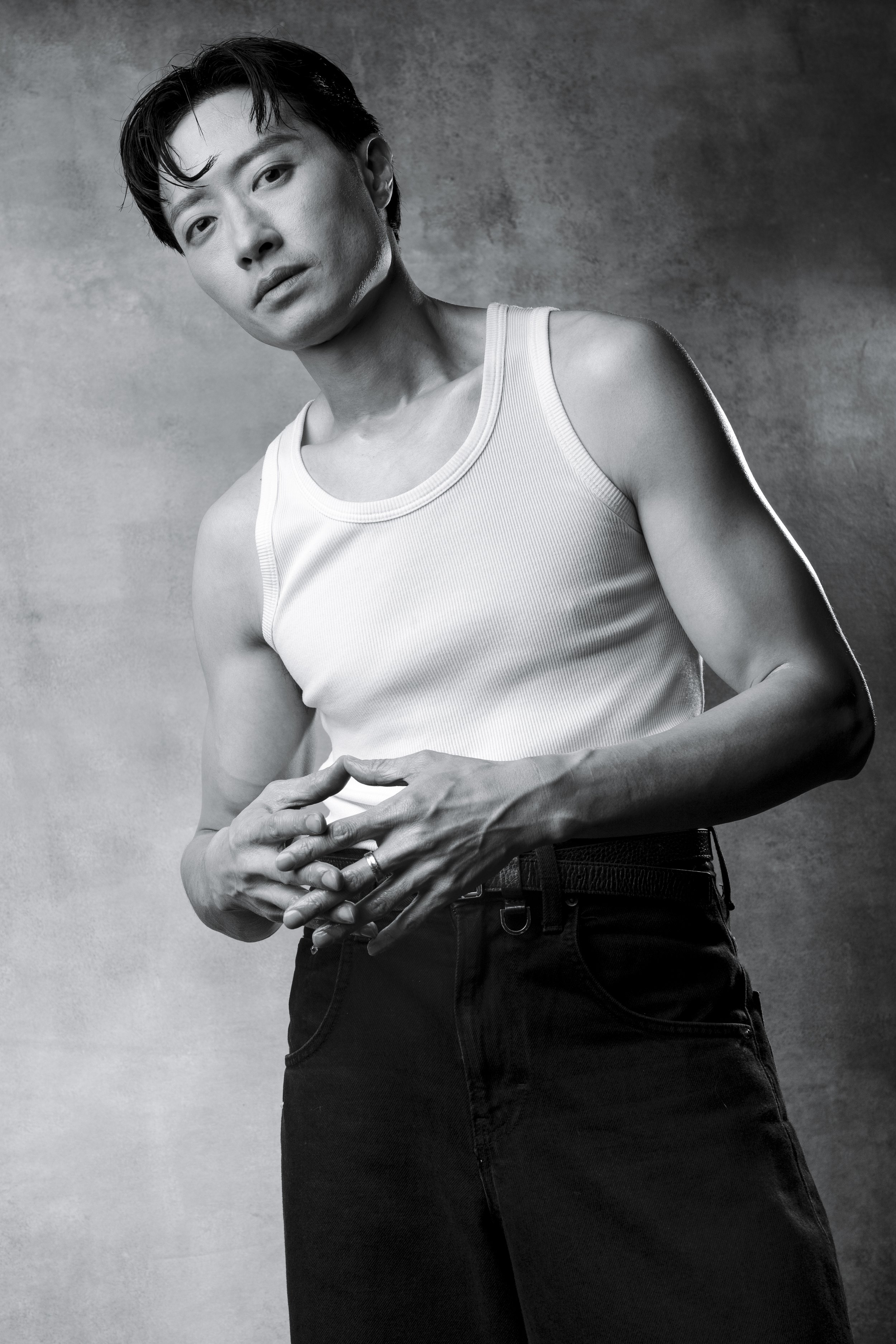 A black-and-white portrait of a person with short dark hair wearing a white tank top and dark pants, standing against a textured background.